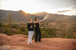 Couple celebrating engagement by popping champagne on red rocks at Garden of the Gods with mountain sunset backdrop.