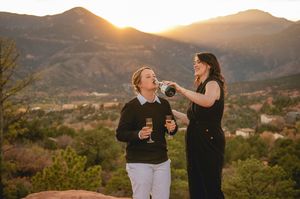 Two women celebrate at sunset in the mountains, one pouring champagne, with scenic Colorado peaks in the background.