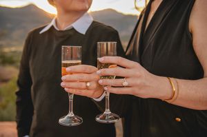 Two women toasting with champagne flutes, wearing engagement rings, at a scenic mountain sunset location.