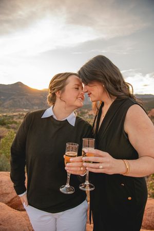 Two women toast with champagne glasses at sunset, touching foreheads romantically with mountain landscape behind them.