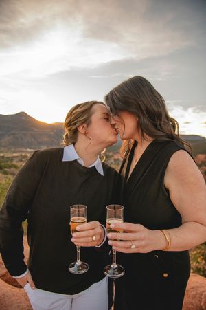 Two women sharing a kiss while holding champagne glasses during a romantic outdoor engagement session at sunset.