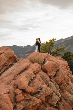 Couple sharing romantic kiss atop red rock formations at Garden of the Gods with mountain views behind them.