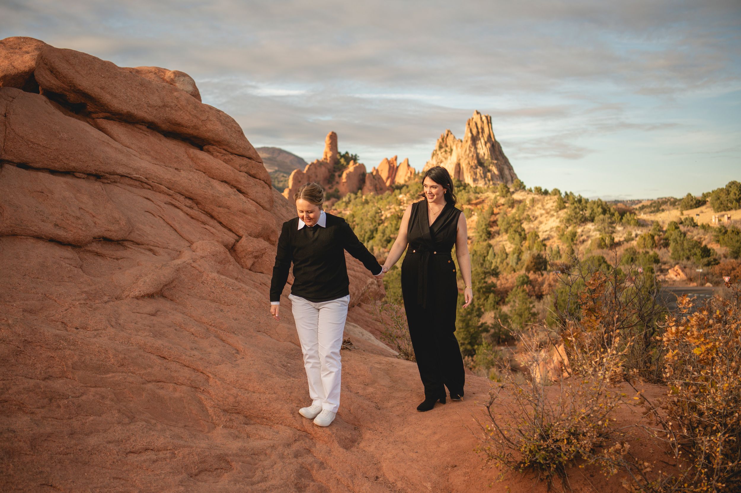 Couple holding hands on red rock trail at Garden of the Gods, Colorado, with dramatic sandstone formations at sunset.