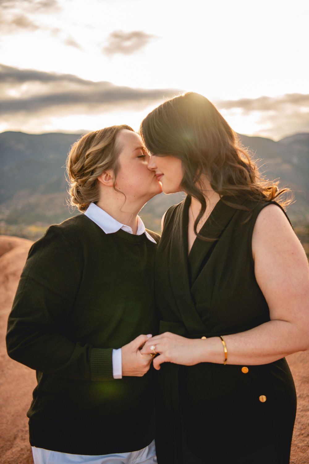 Two women sharing a romantic kiss at golden hour with mountains and dramatic sky in the background.