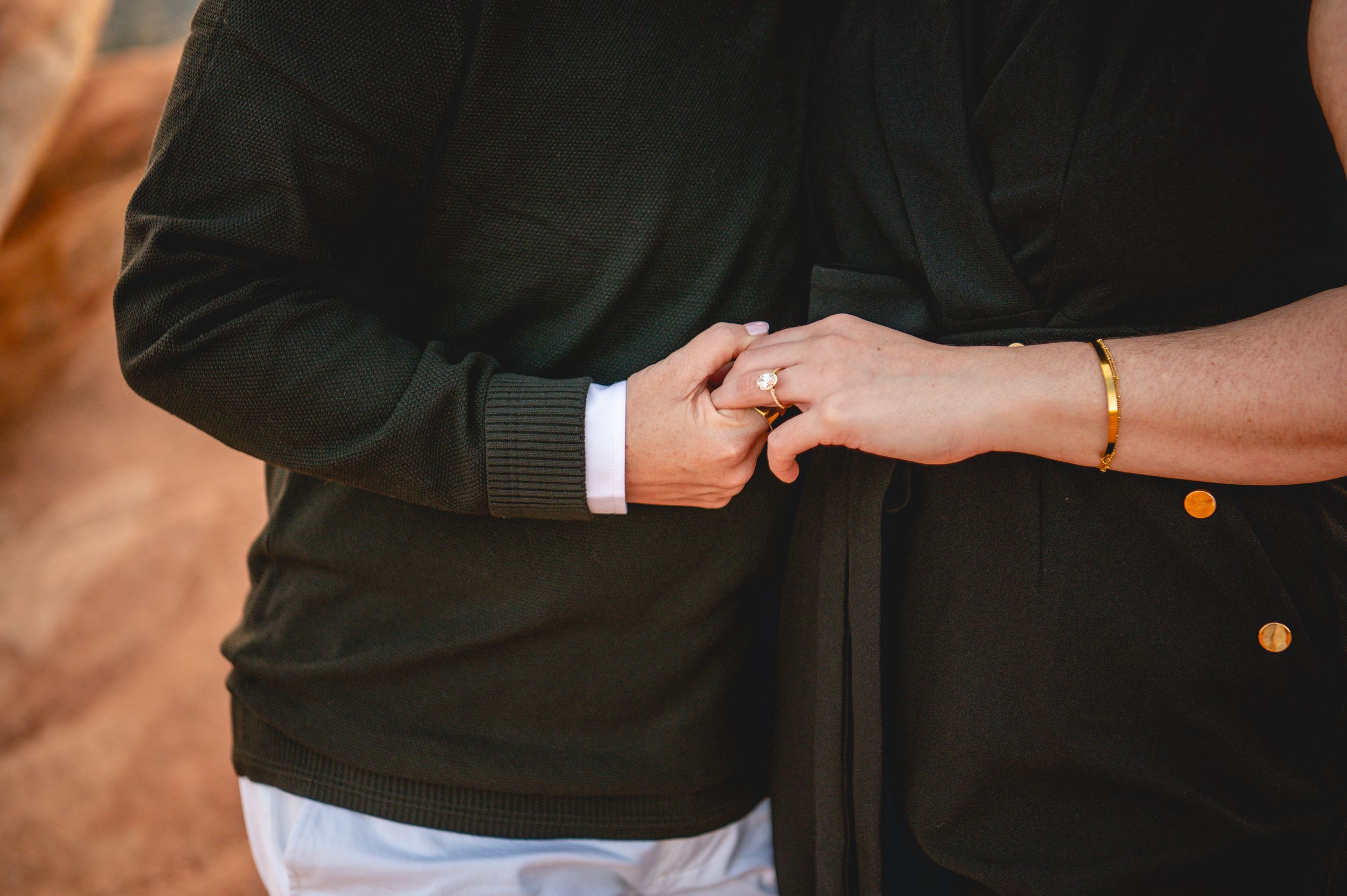 Couple holding hands closely, wearing dark green and black outfits, with a gold ring and bracelet visible.