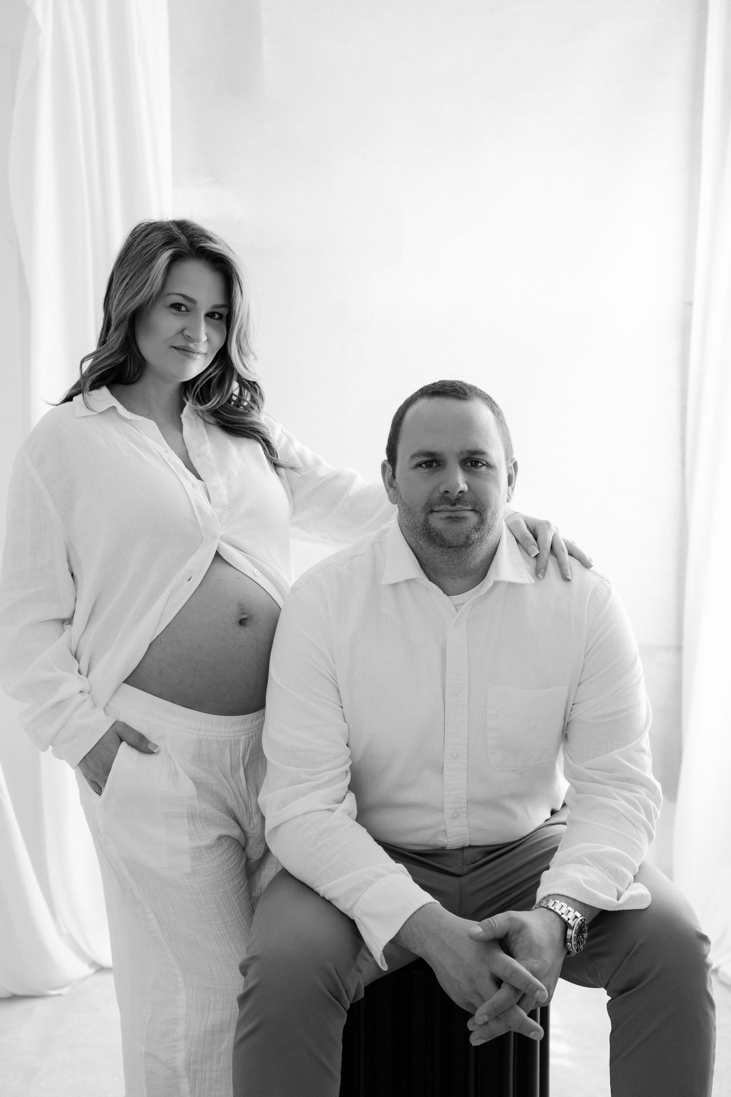 Black and white maternity photo of pregnant woman in white shirt with partner seated beside her in bright studio.