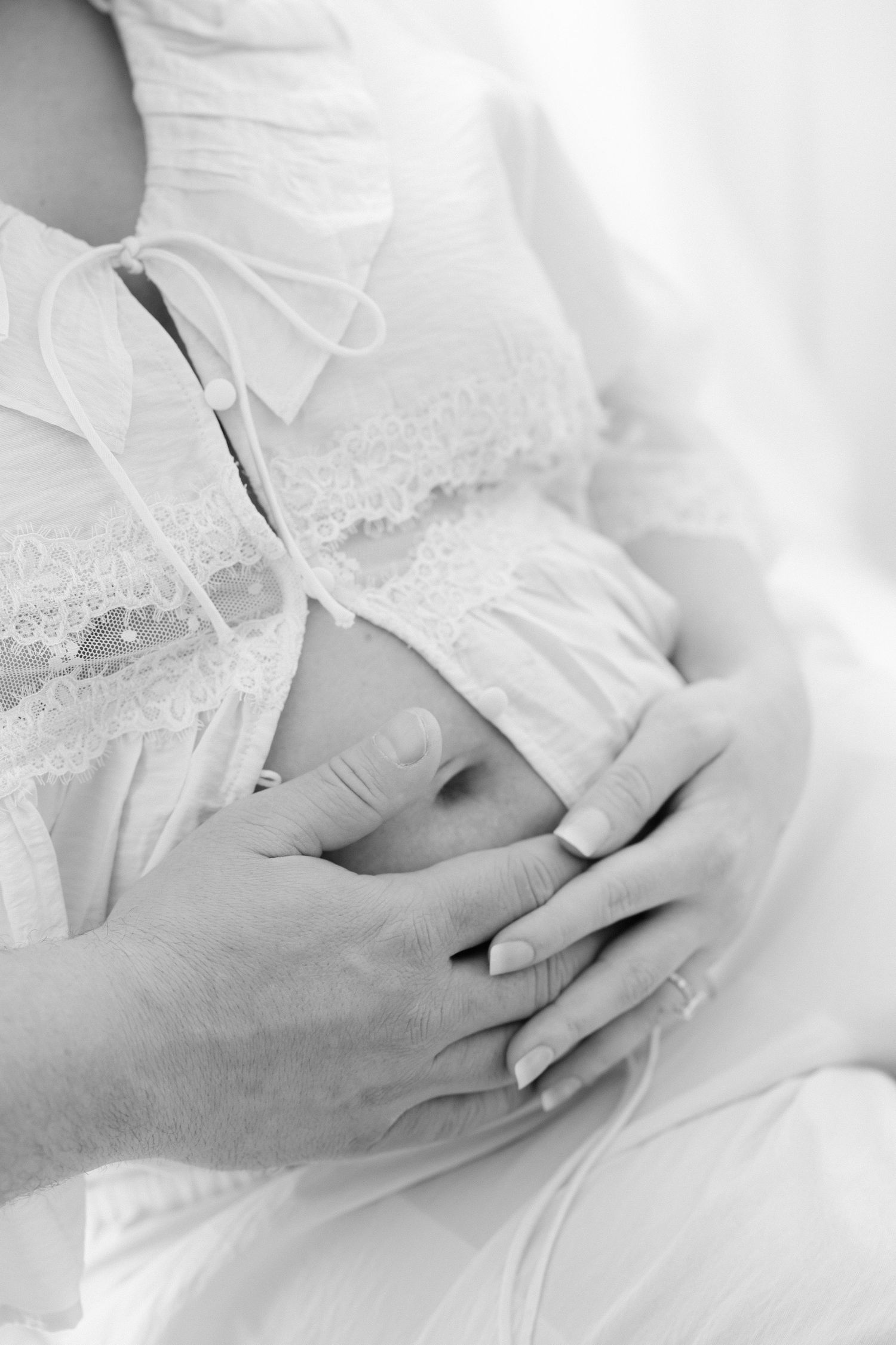 Black and white photo of a pregnant woman in white lace top, cradling her baby bump with both hands.