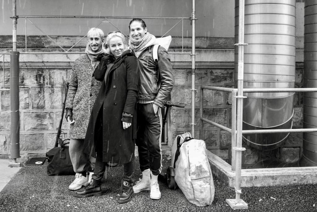 Three friends pose together outdoors in black and white photo, wearing winter coats with luggage nearby.