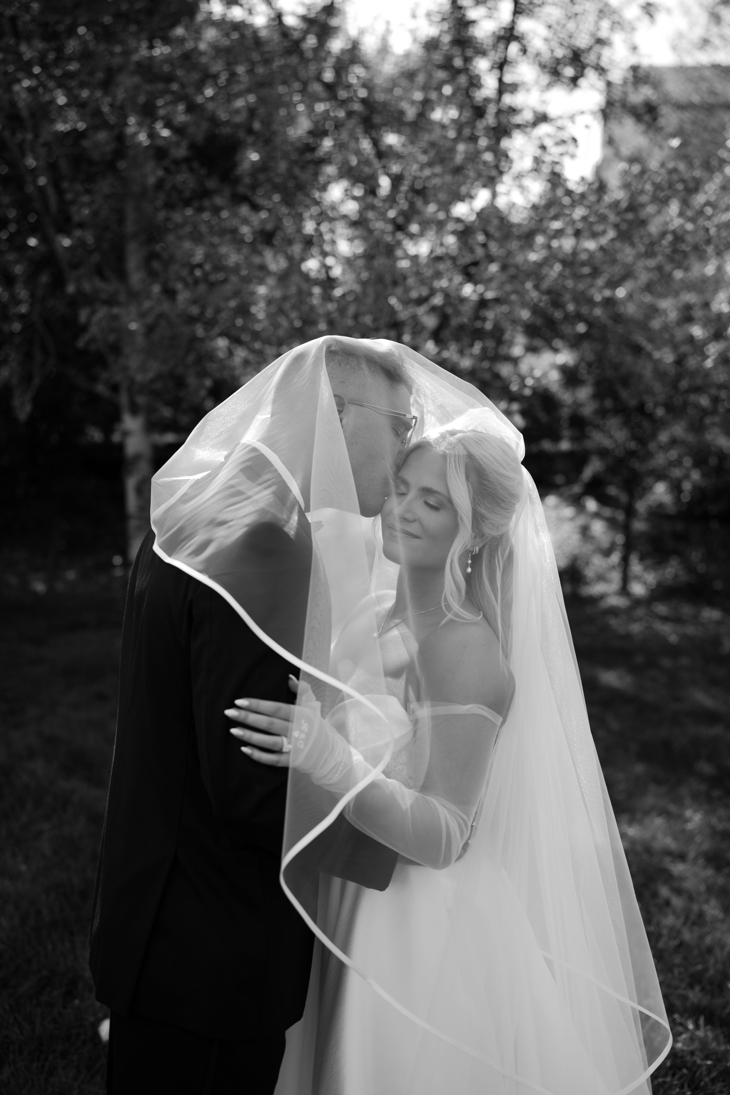 Black and white photo of bride and groom sharing intimate moment under wedding veil outdoors.