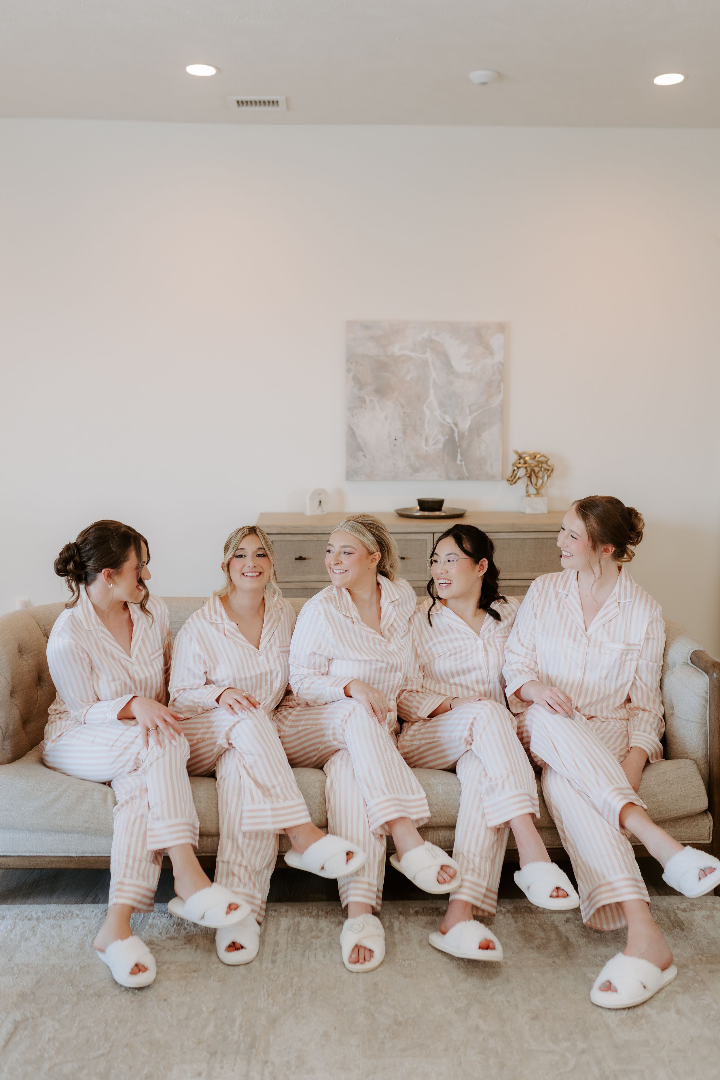 Five bridesmaids in matching pink striped pajamas and white slippers sitting together on a couch laughing during bridal party preparations.