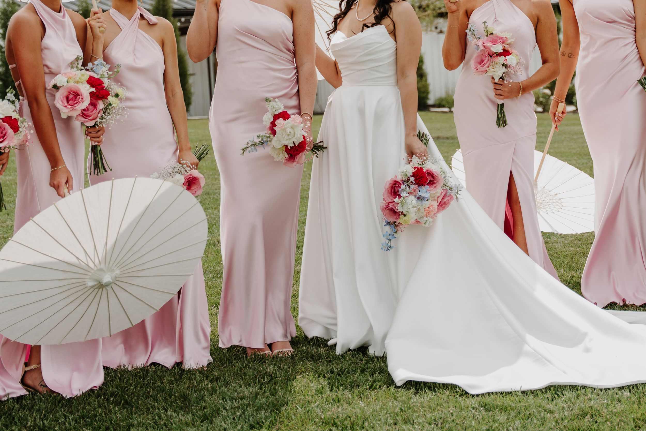 Bride in white gown with bridesmaids in pink dresses holding floral bouquets and white parasols outdoors.