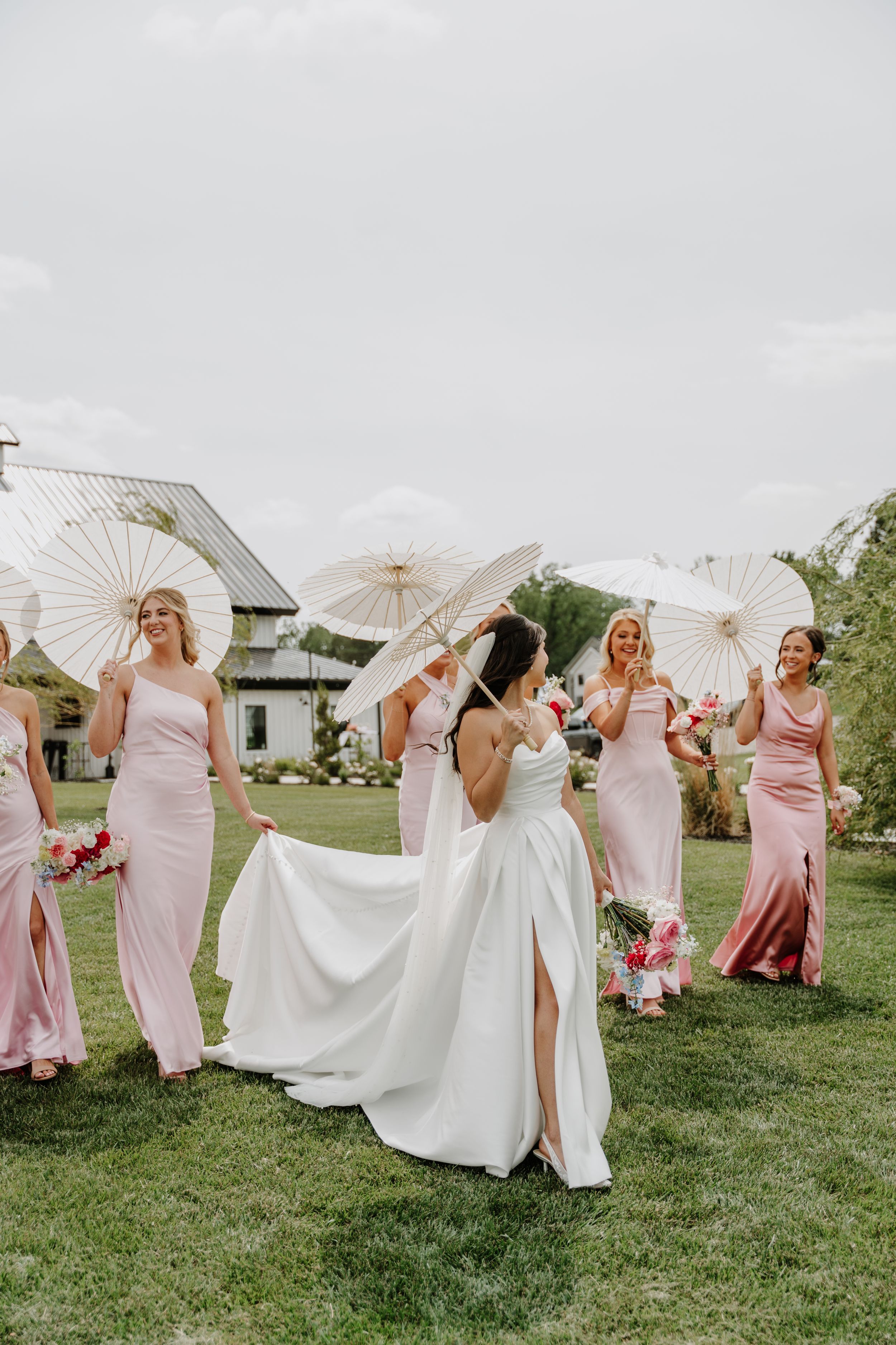 Bride in white gown holds parasol with bridesmaids in pink dresses carrying white parasols on green lawn.