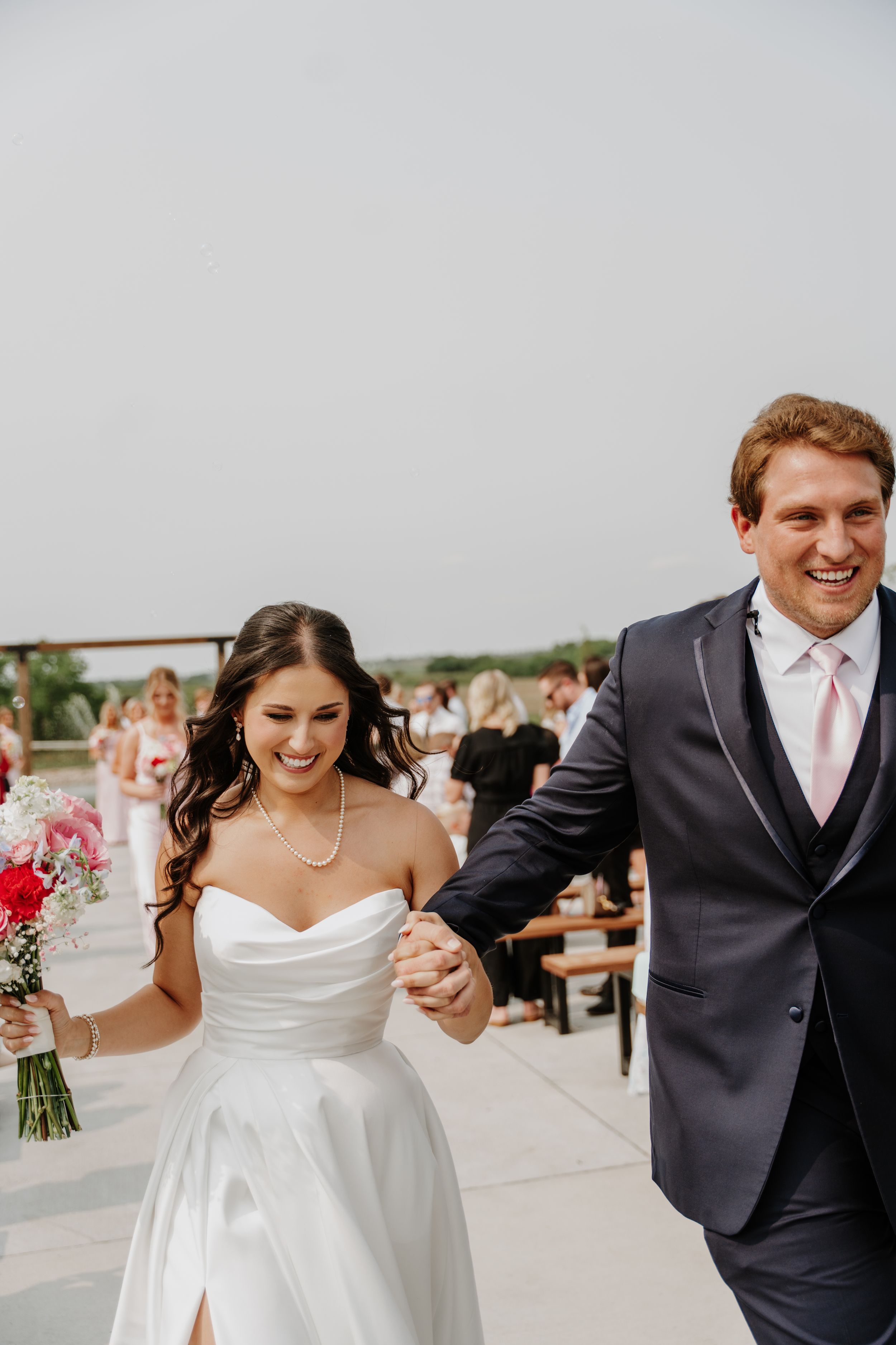 Joyful newlyweds walk hand in hand after outdoor wedding ceremony, bride in white gown holding pink bouquet, groom in navy suit.