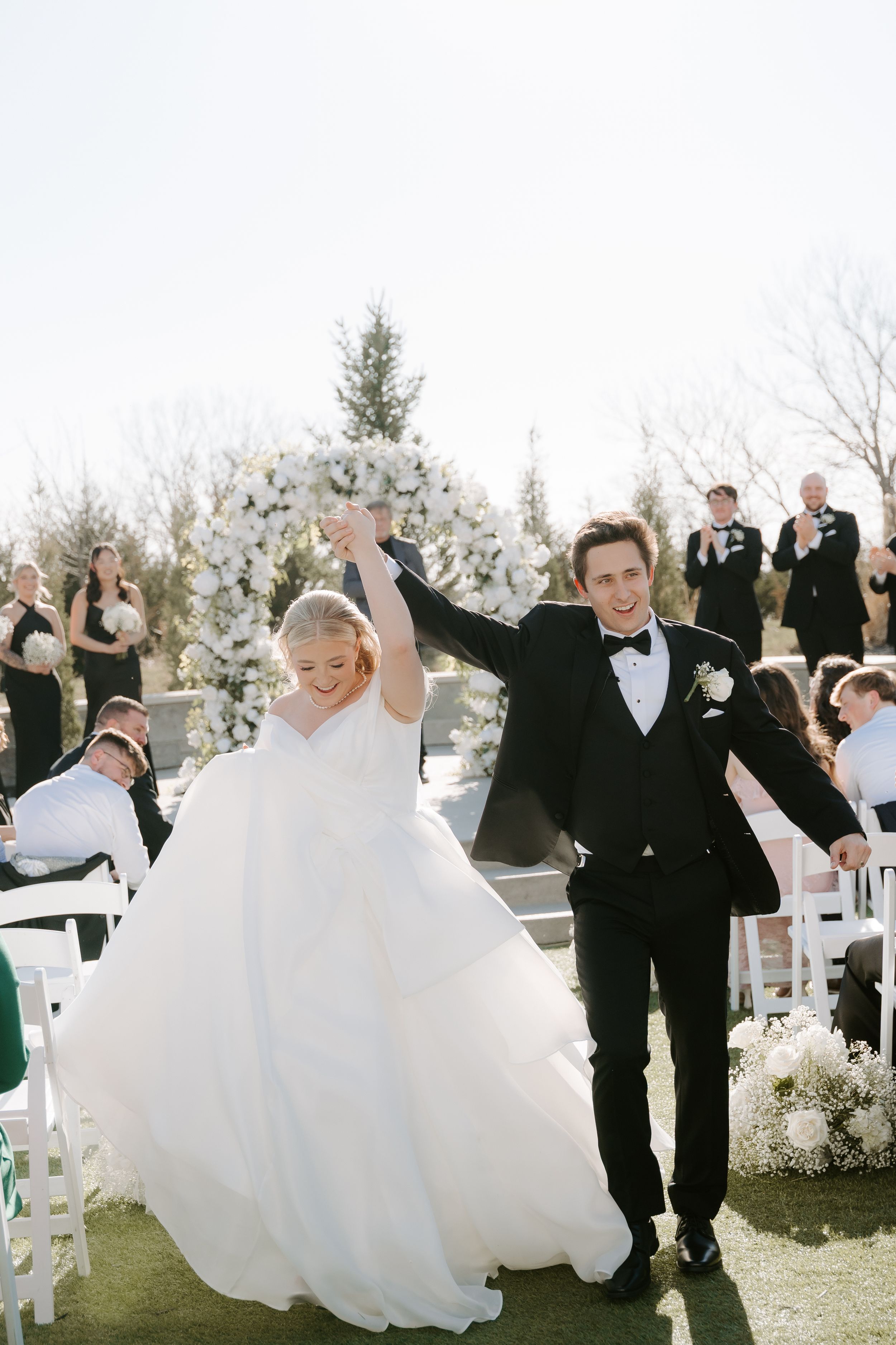 Joyful newlyweds raise hands walking down outdoor wedding aisle, bride in white gown holding bouquet, groom in black tuxedo.