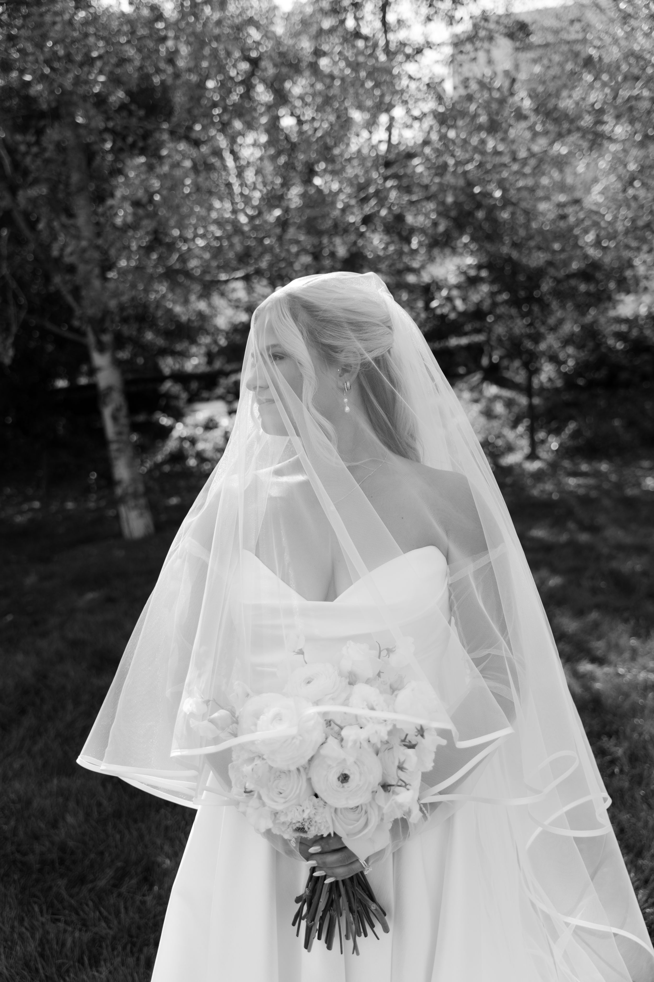 Black and white photo of a bride in a strapless gown, veil draped over her head, holding a bouquet outdoors.