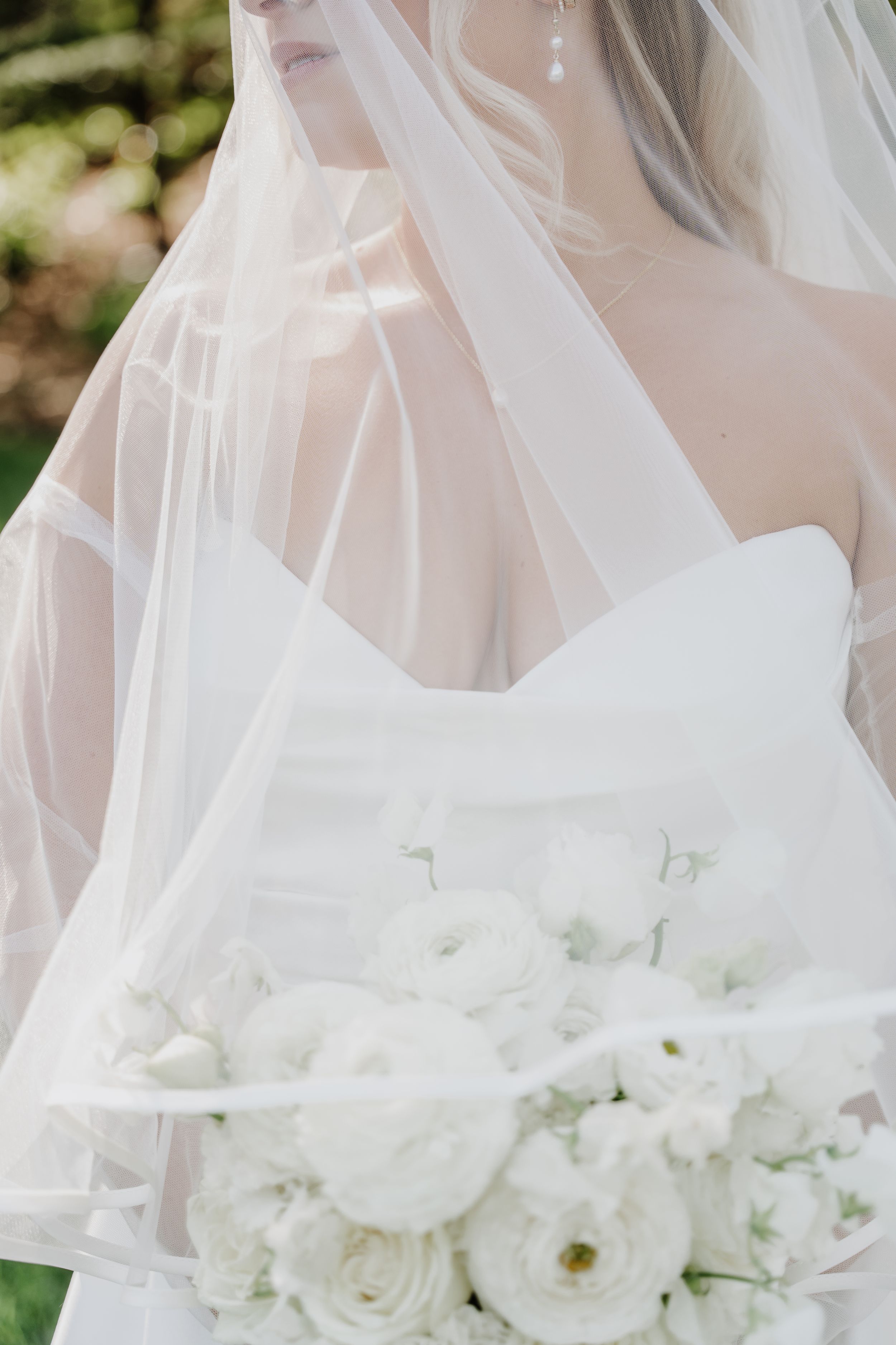 Bride in white strapless gown holding elegant all-white floral bouquet, draped in sheer veil, soft natural light.