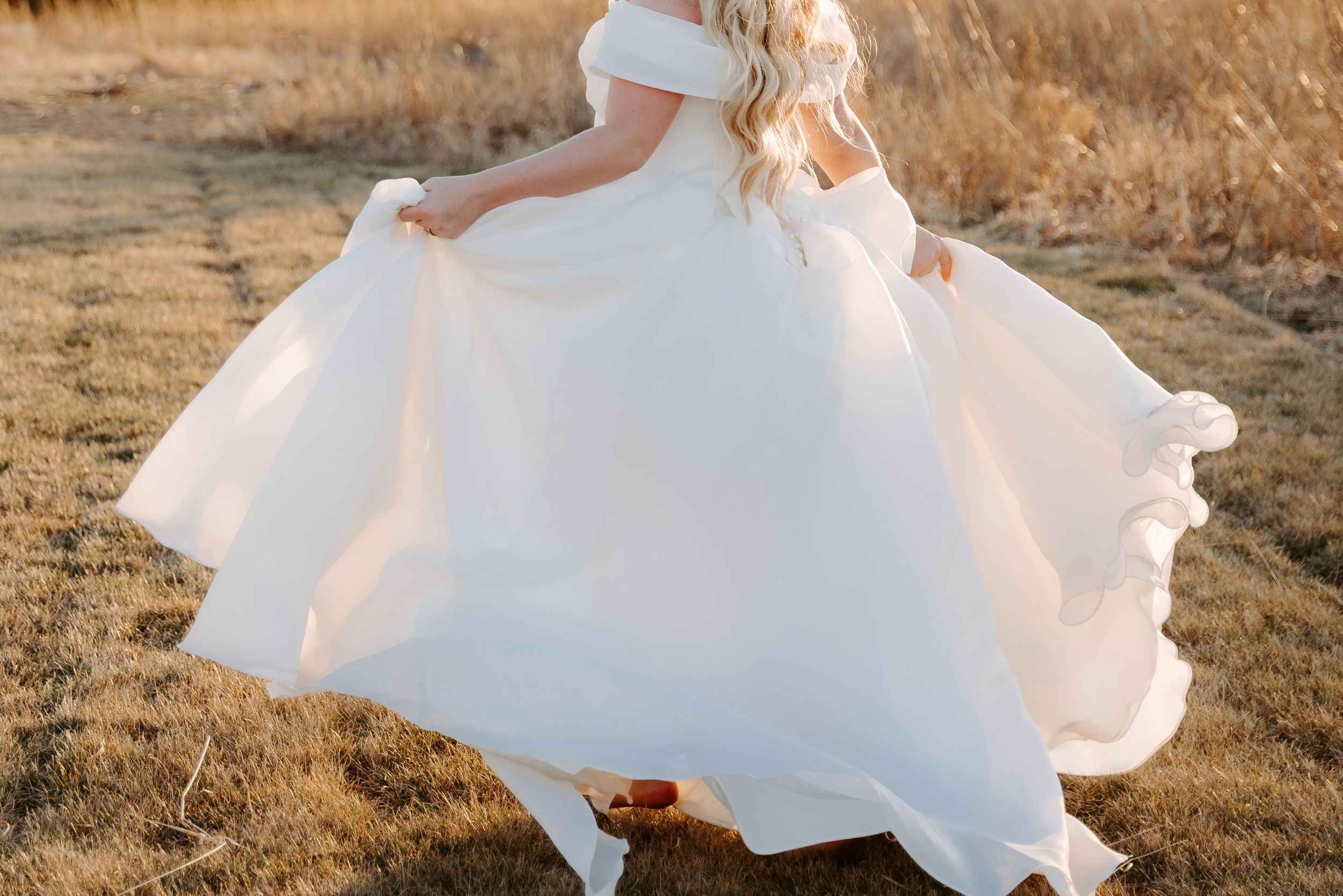 A bride twirls in a flowing white off-shoulder wedding gown in a golden field during sunset.