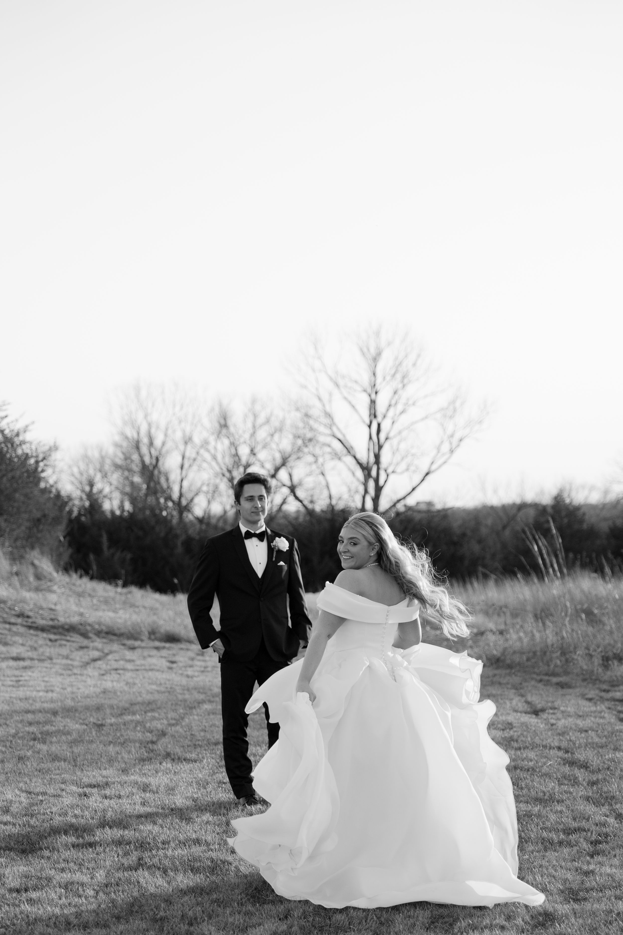 Black and white photo of bride in off-shoulder gown and groom in tuxedo walking through a field.