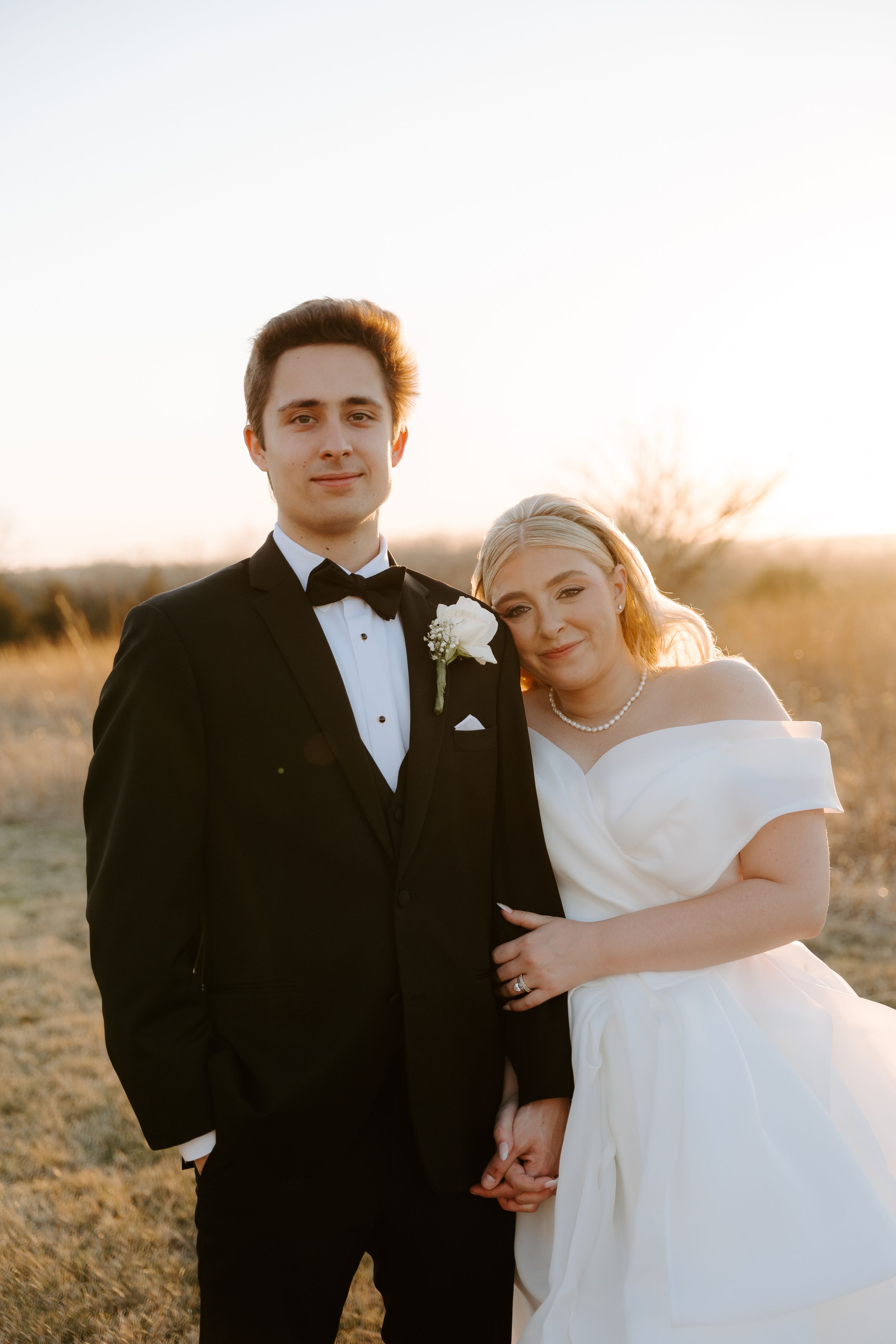 Bride and groom posing together at golden hour in a field, she in white off-shoulder gown, he in classic black tuxedo.