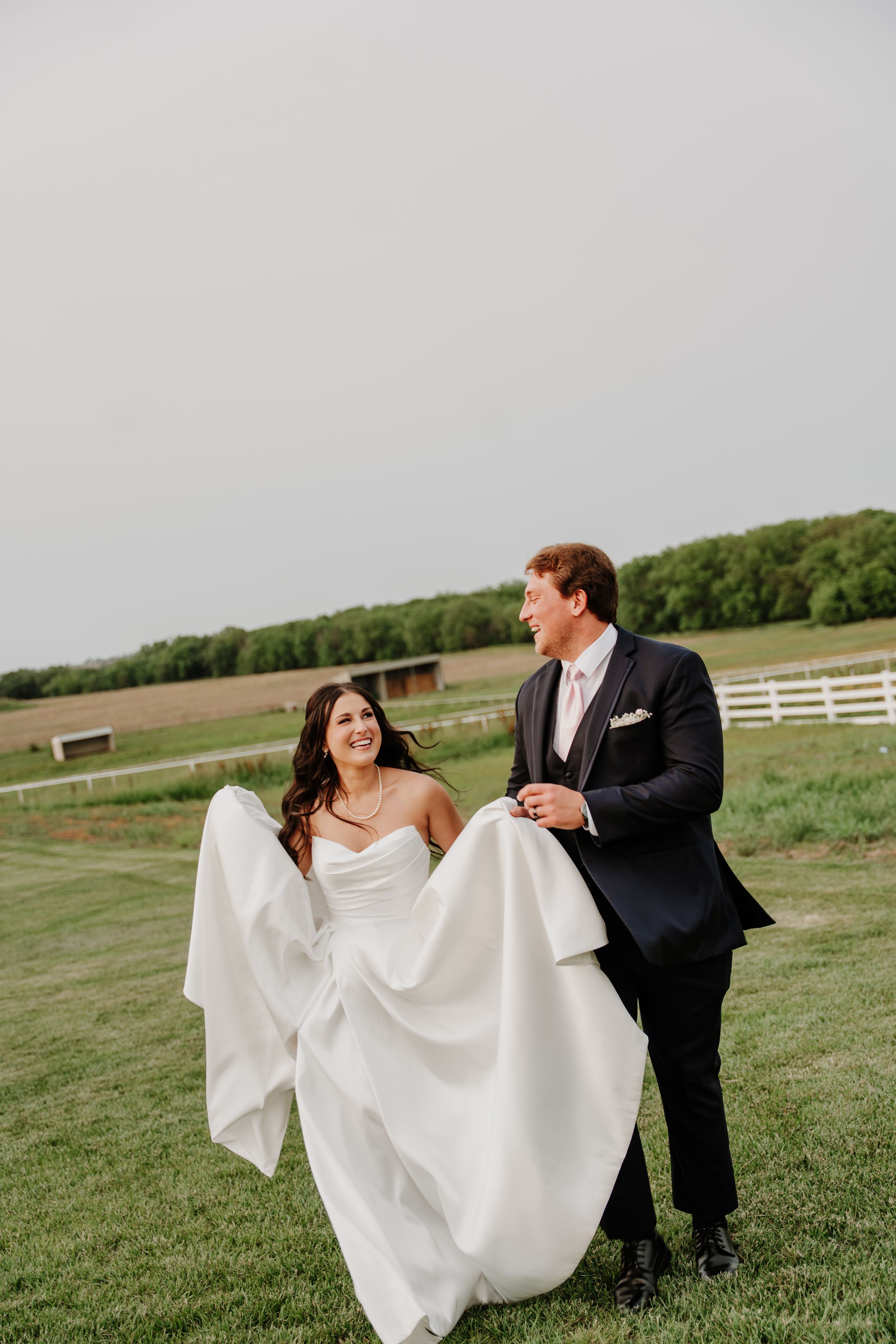 A smiling bride and groom walk hand-in-hand across a green lawn at a rustic farm wedding venue.