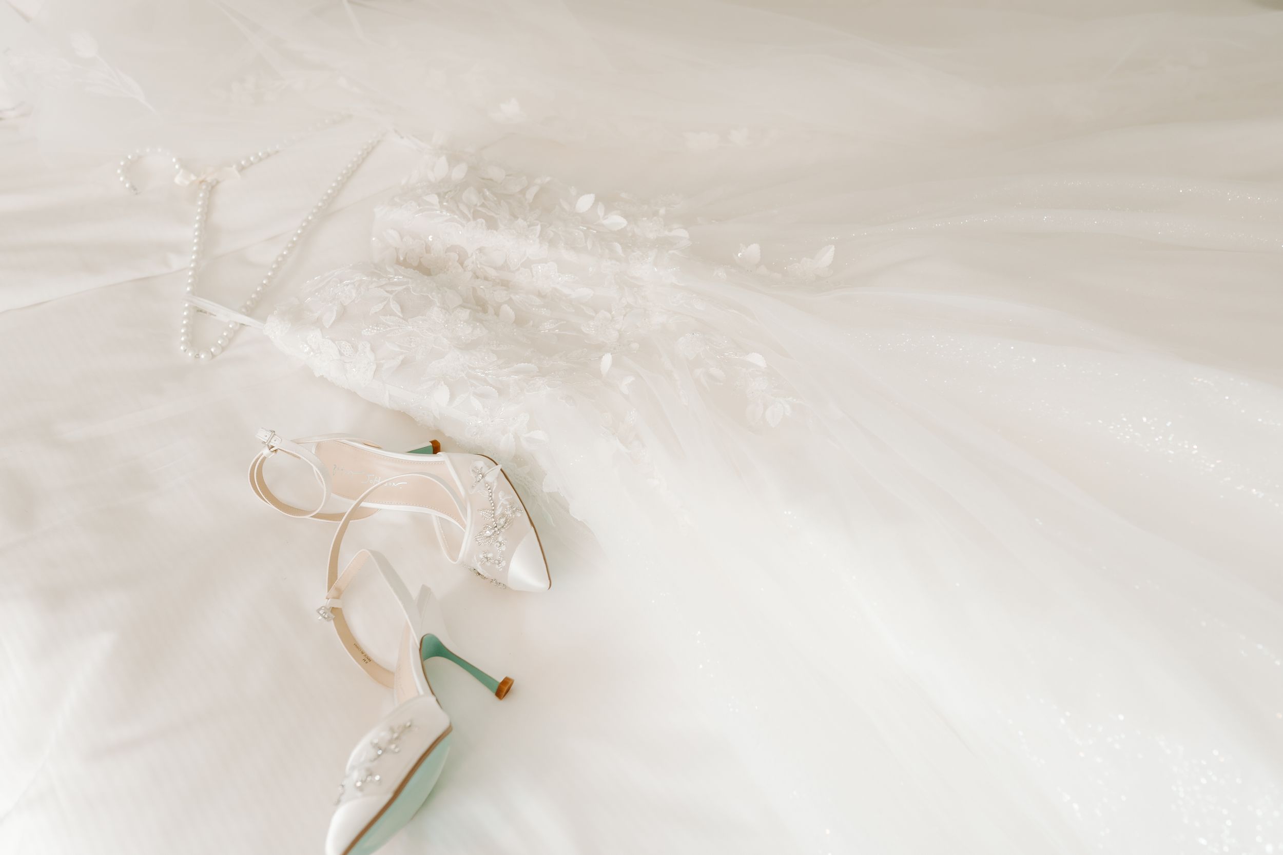 Elegant white bridal heels with ankle straps resting on a flowing wedding dress with lace details.