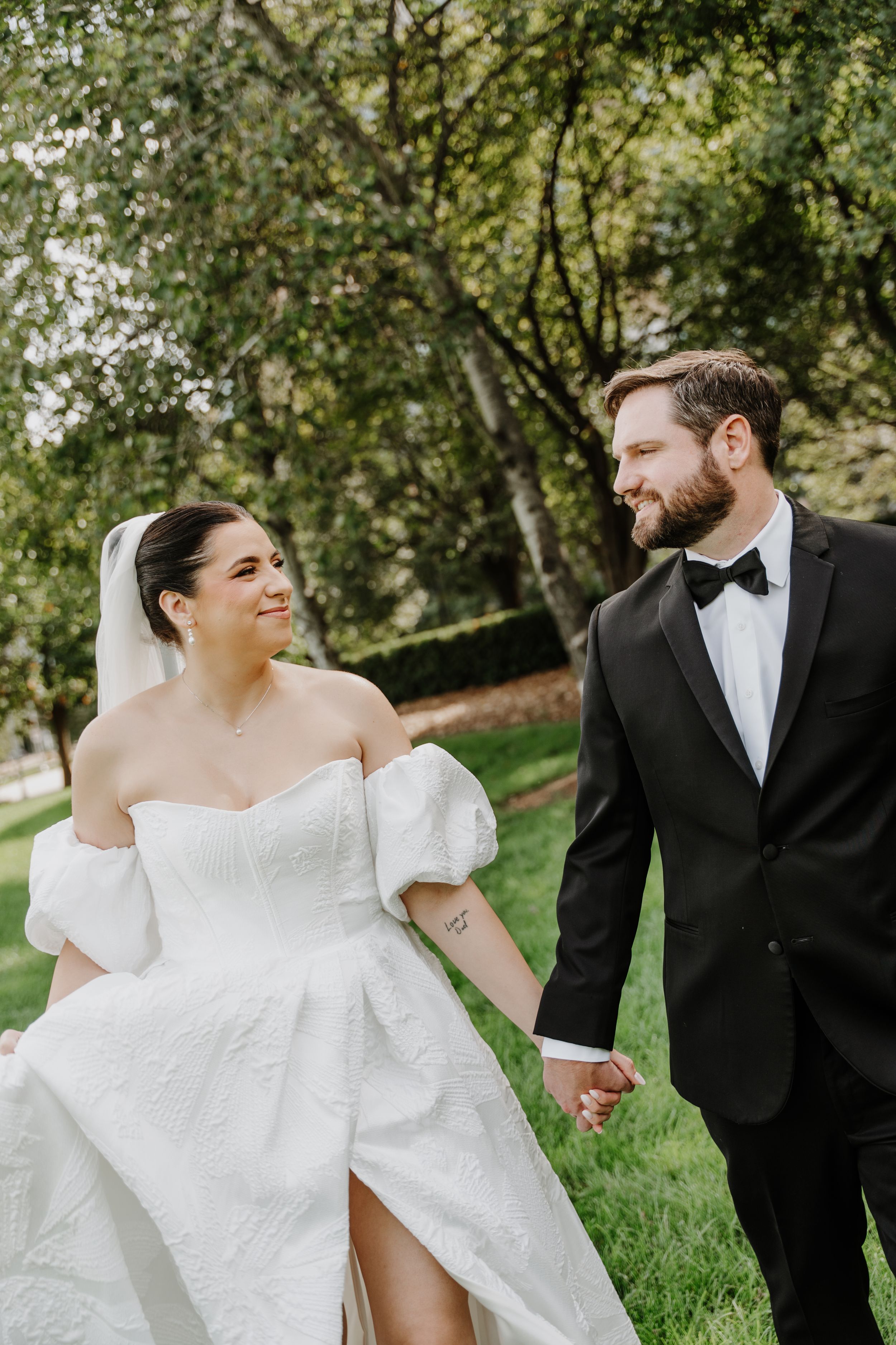 A bride in a white off-shoulder gown and groom in black tuxedo hold hands walking through a lush green park.