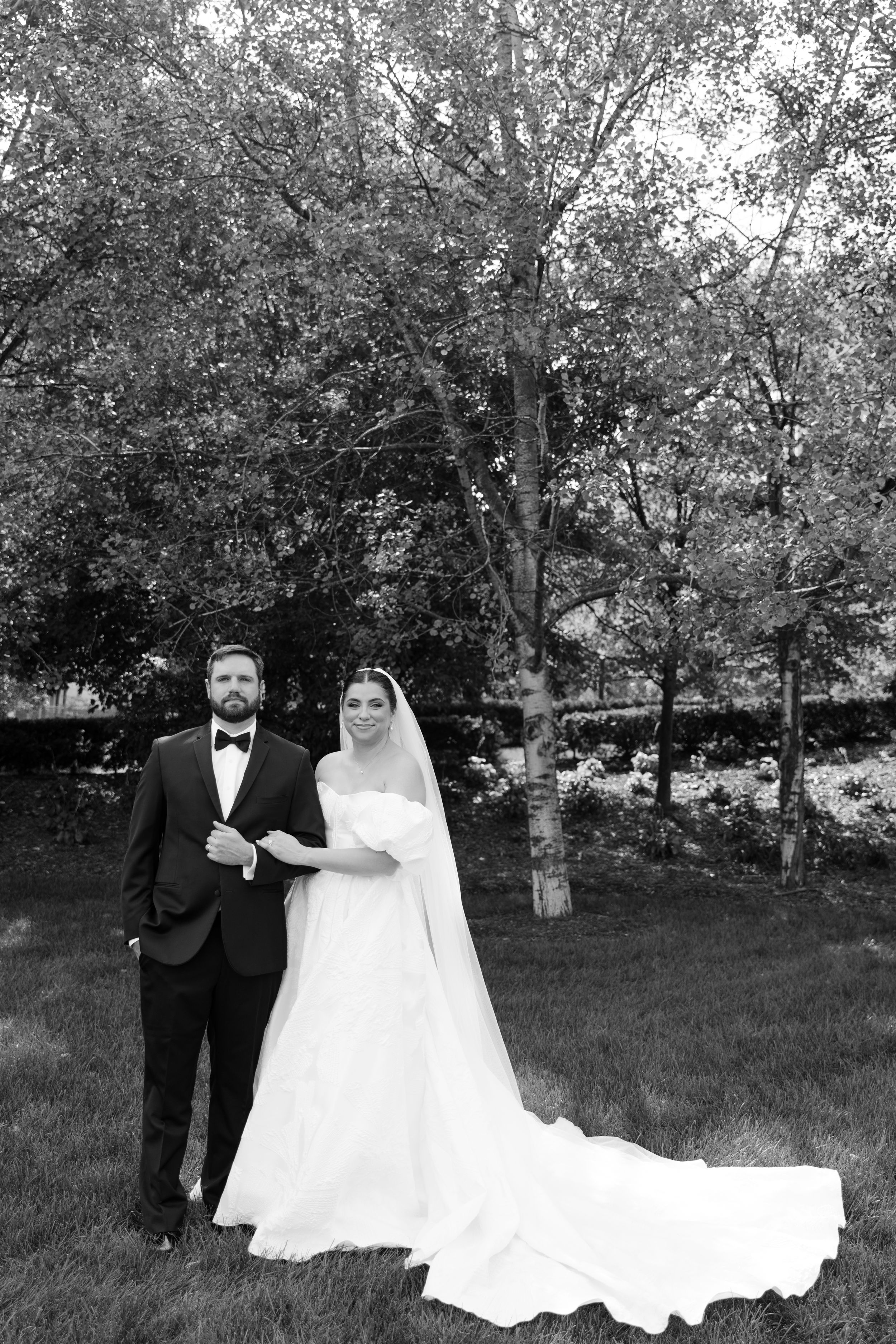 Black and white wedding portrait of bride in flowing gown with veil and groom in tuxedo standing outdoors among trees.