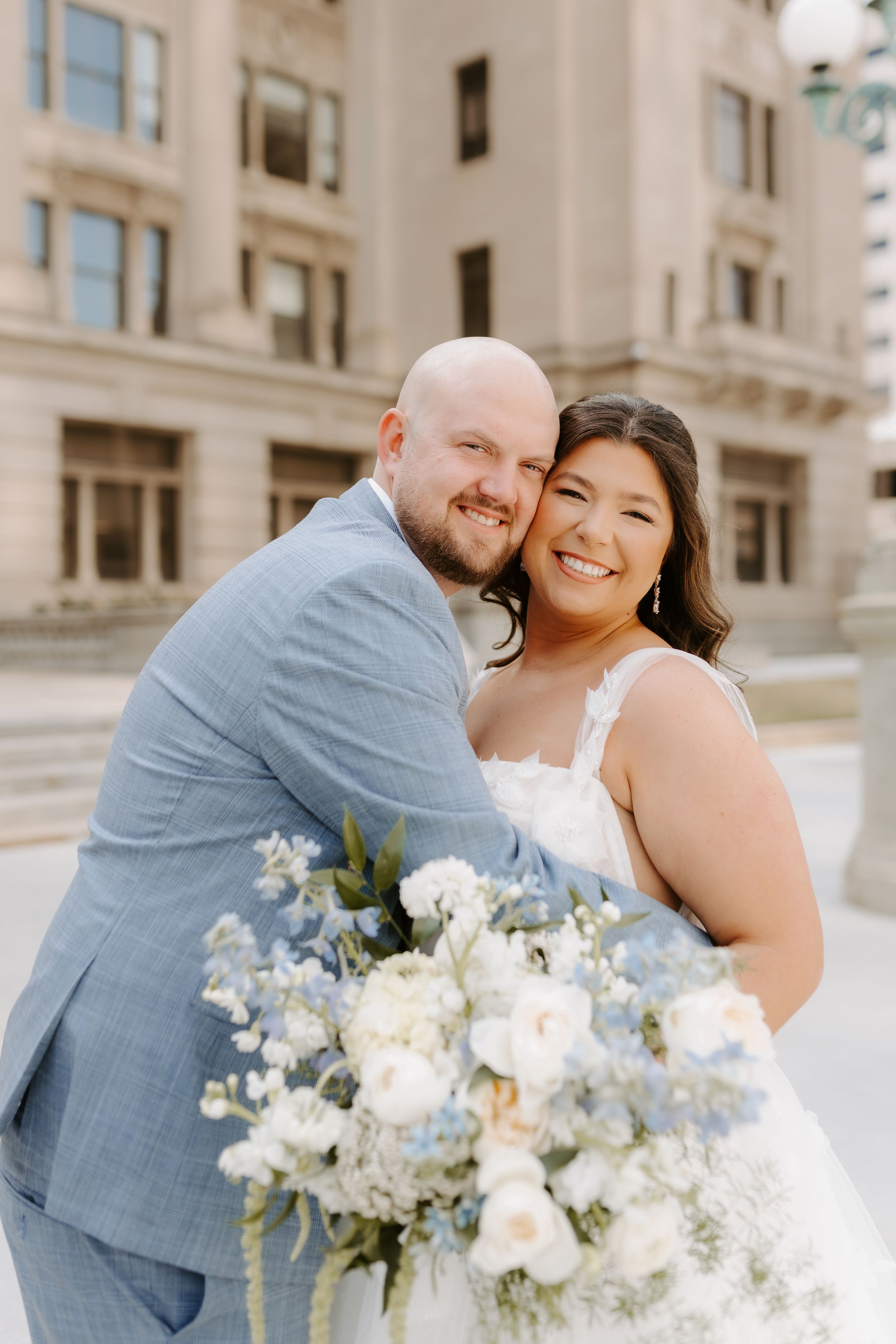 A smiling bride and groom embrace outdoors, she holds a blue and white floral bouquet, he wears a light blue suit.