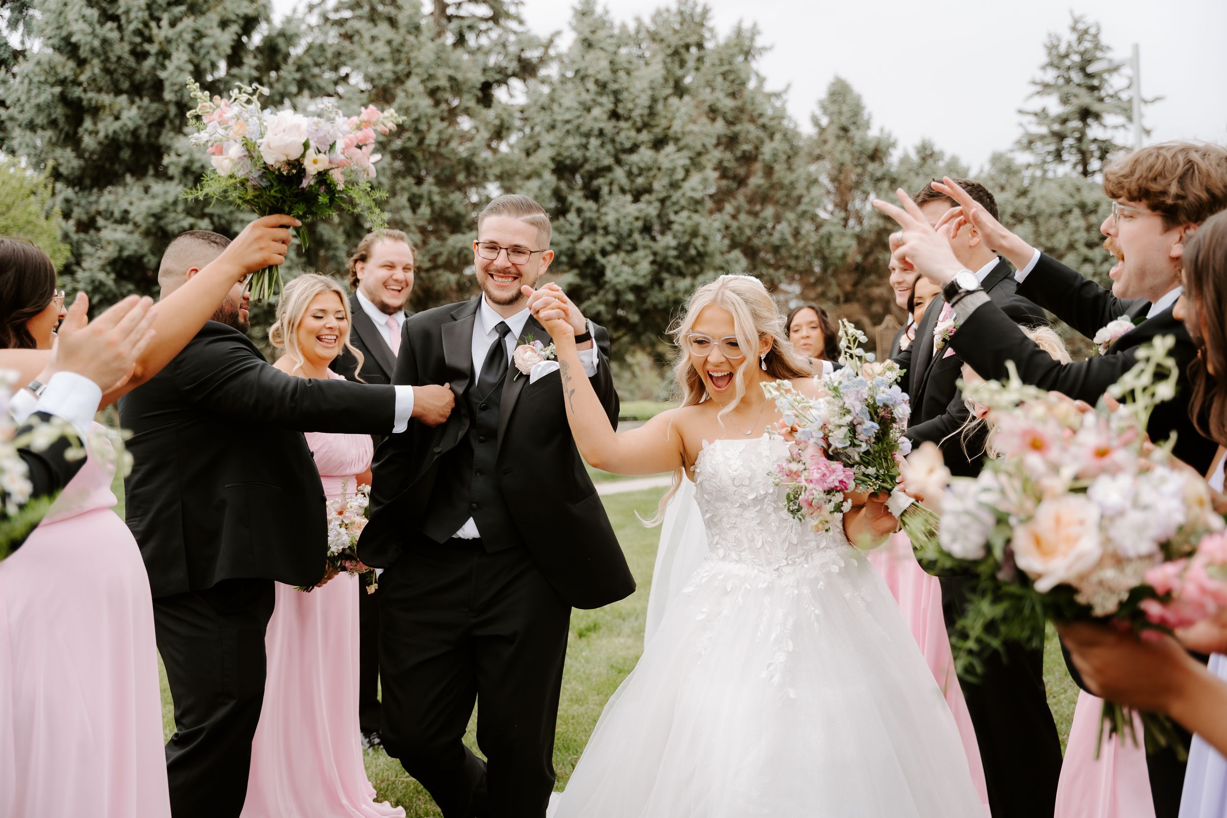 Bride and groom walk joyfully through cheering wedding party in pink dresses and black tuxedos outdoors.