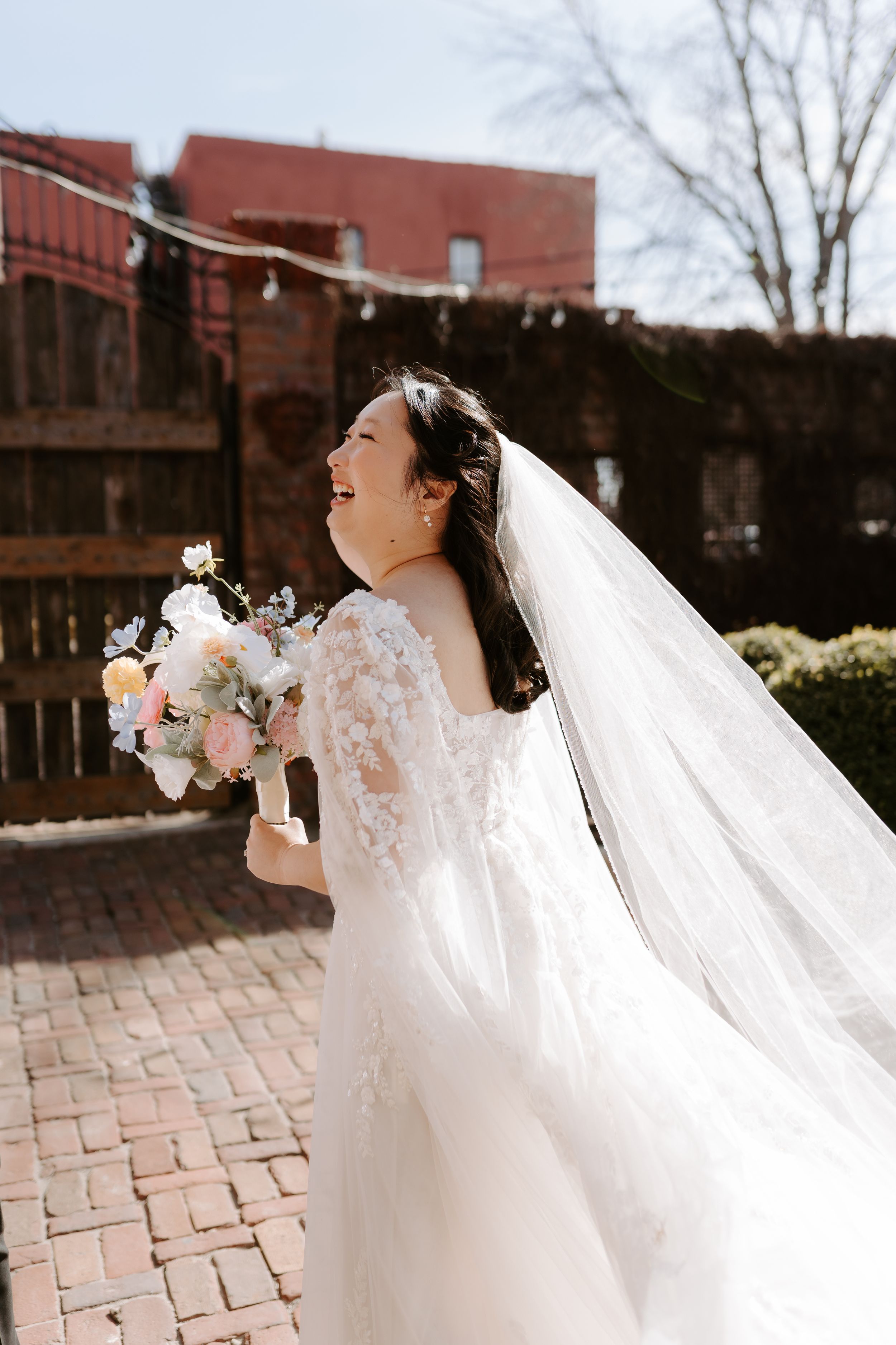 Joyful bride in lace off-shoulder gown and flowing veil holds pastel bouquet, laughing outdoors on brick patio.