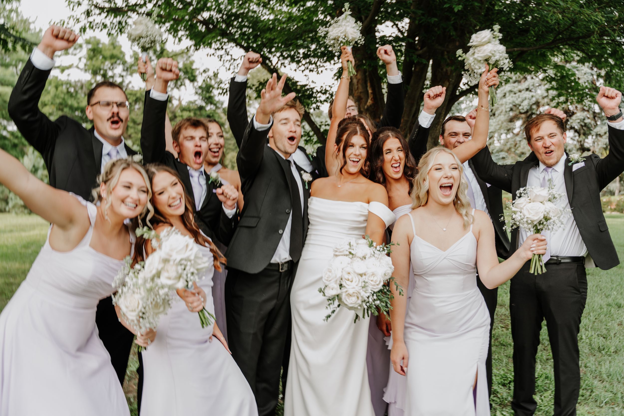 Joyful wedding party celebrating outdoors, bride in white gown holding bouquet, bridesmaids and groomsmen cheering with raised arms.