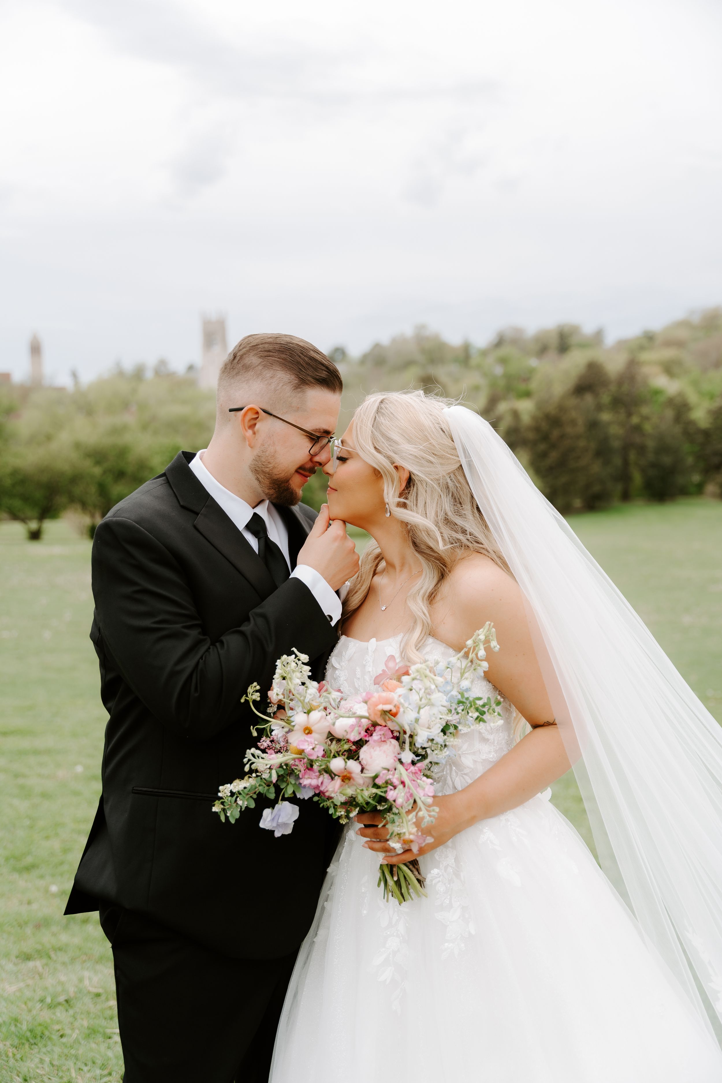 Bride and groom share a romantic moment outdoors, bride holding a pink floral bouquet in a white gown with veil.