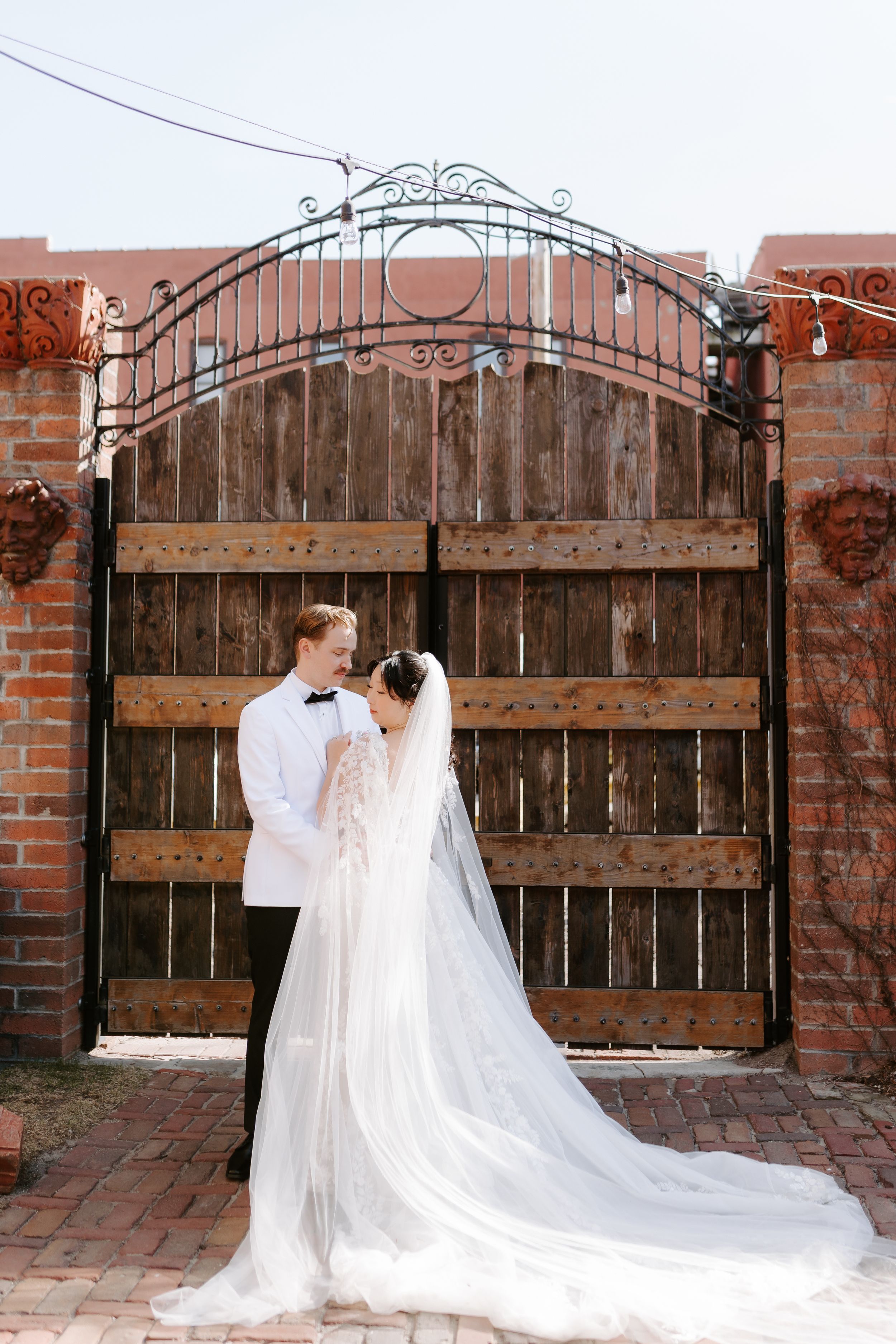 Bride in flowing white gown and veil stands with groom in white tuxedo before rustic wooden gate and brick wall.