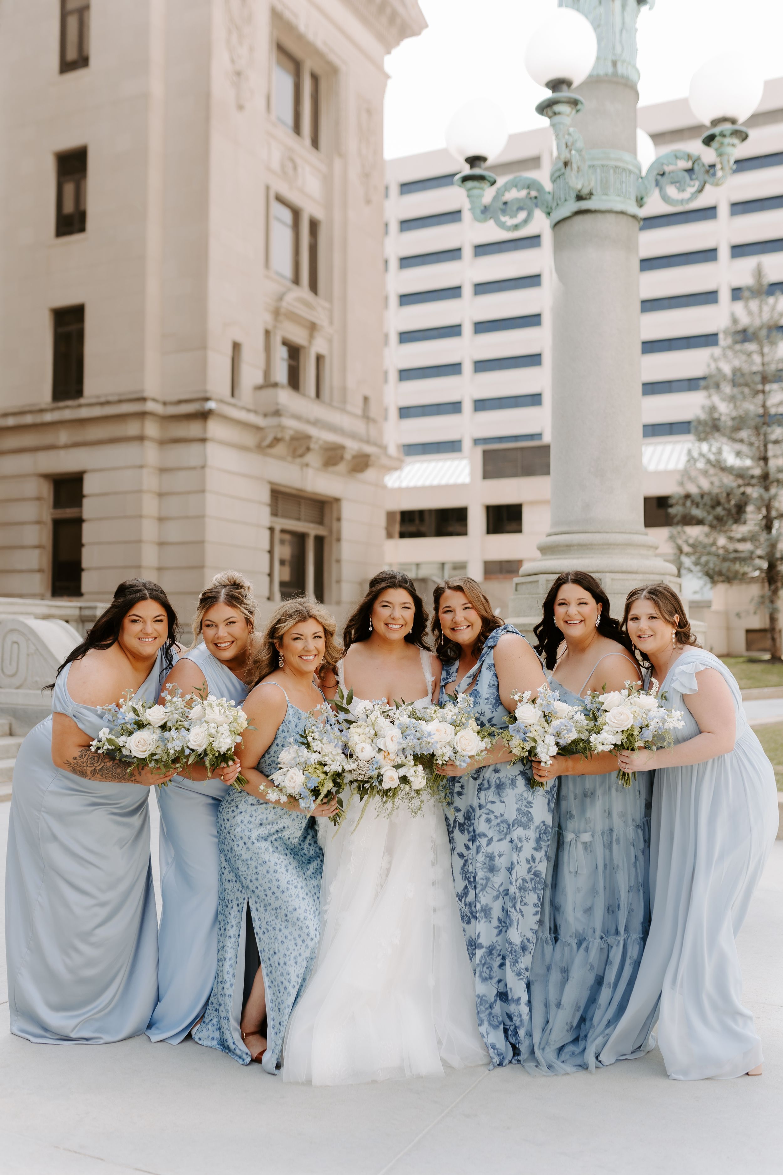 Bride in white gown poses with six bridesmaids in blue dresses holding white bouquets outside a historic building.
