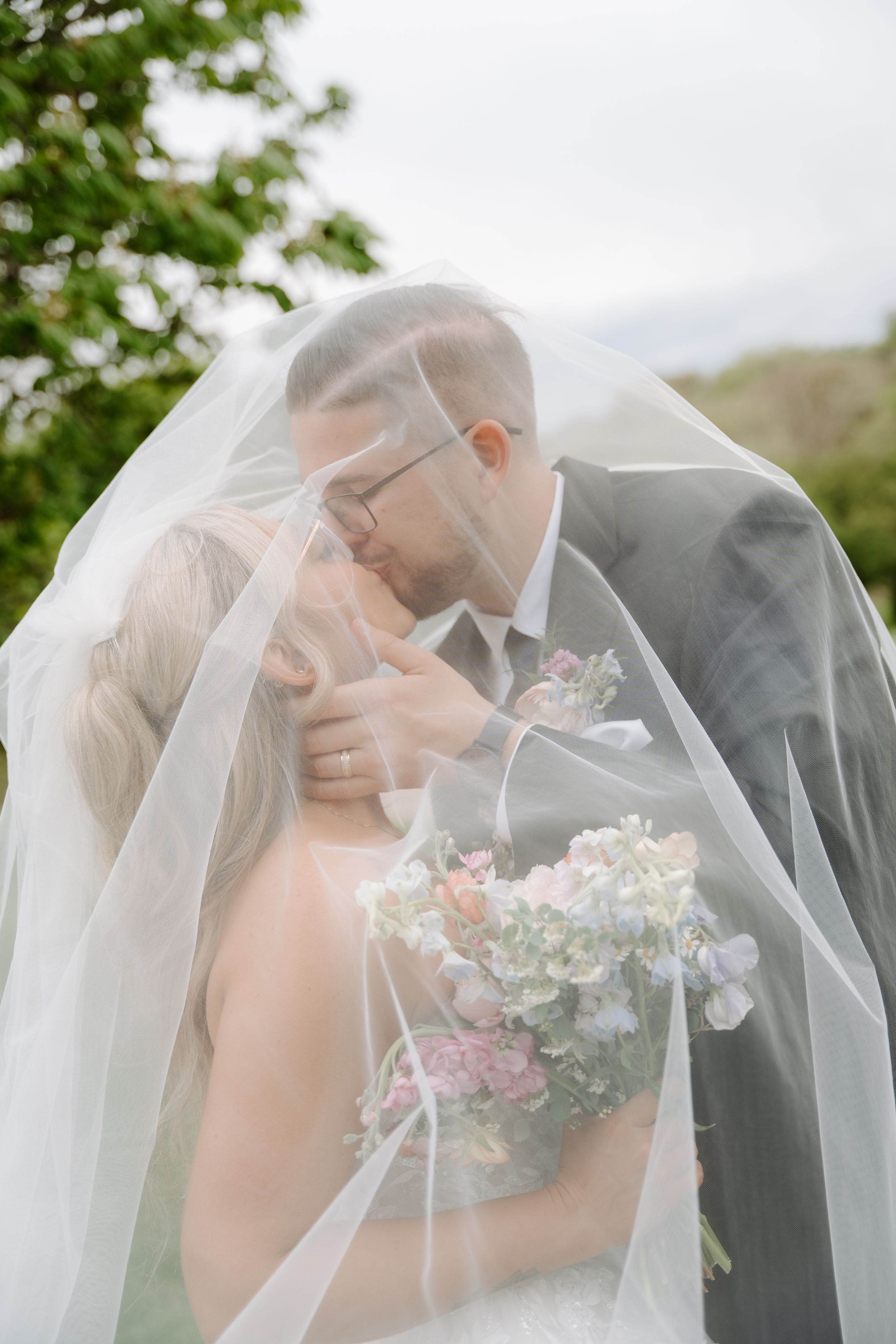 Bride and groom sharing a romantic kiss under a flowing white veil, bride holding a pastel floral bouquet outdoors.