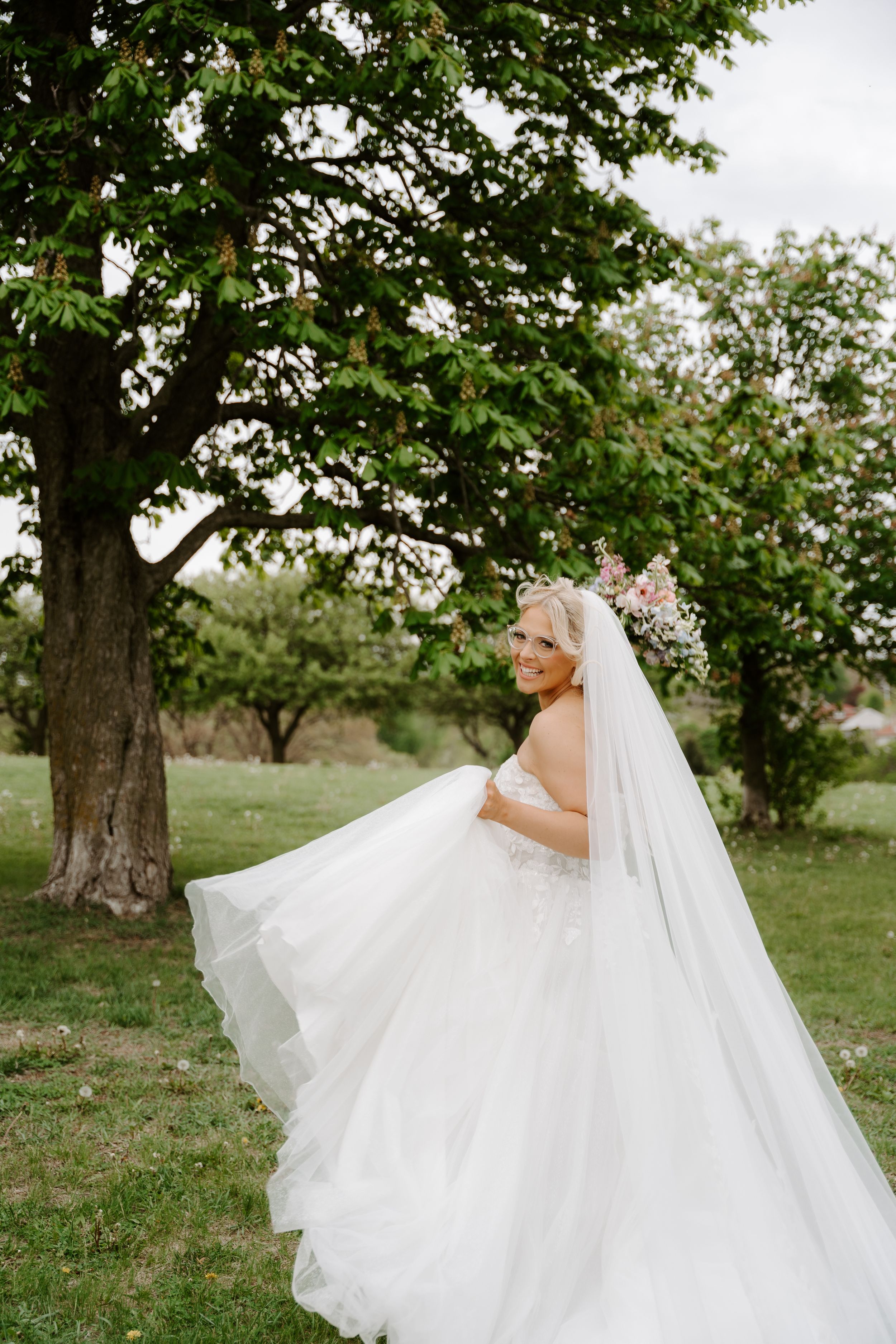Smiling bride in white ballgown and veil poses outdoors near large green tree on wedding day.