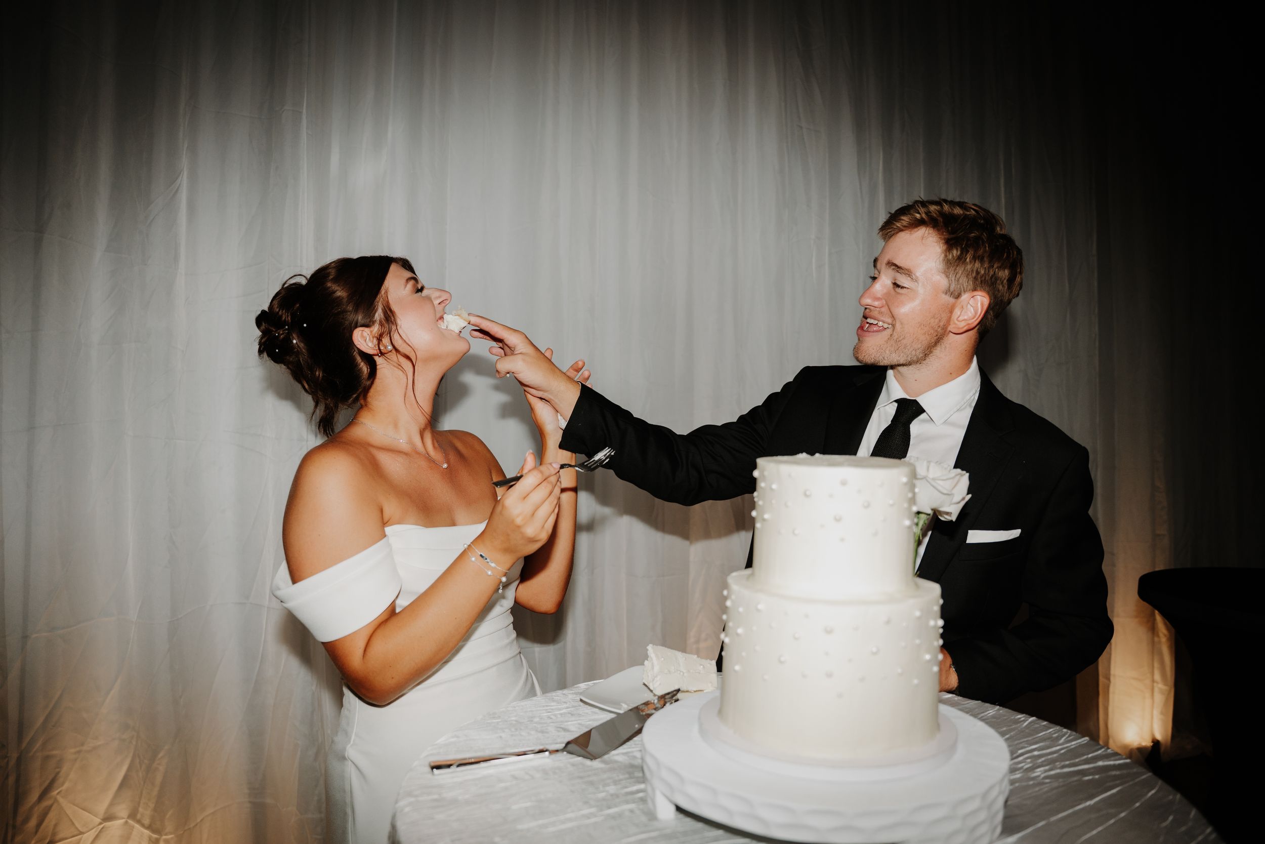 Bride and groom smiling while feeding each other white wedding cake at their reception celebration.