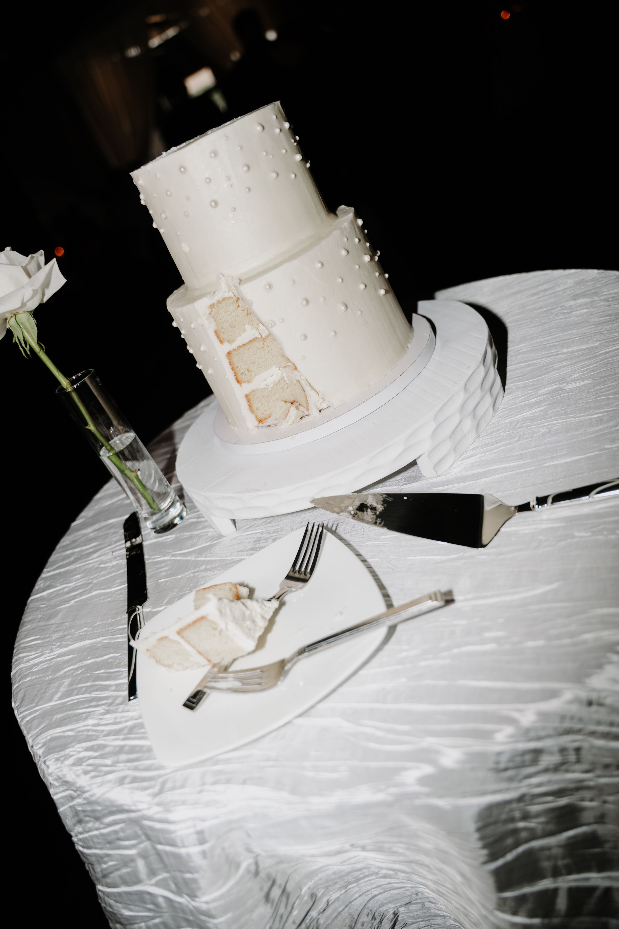 Elegant two-tier white wedding cake with pearl details, partially sliced, displayed on a white tablecloth with serving utensils.