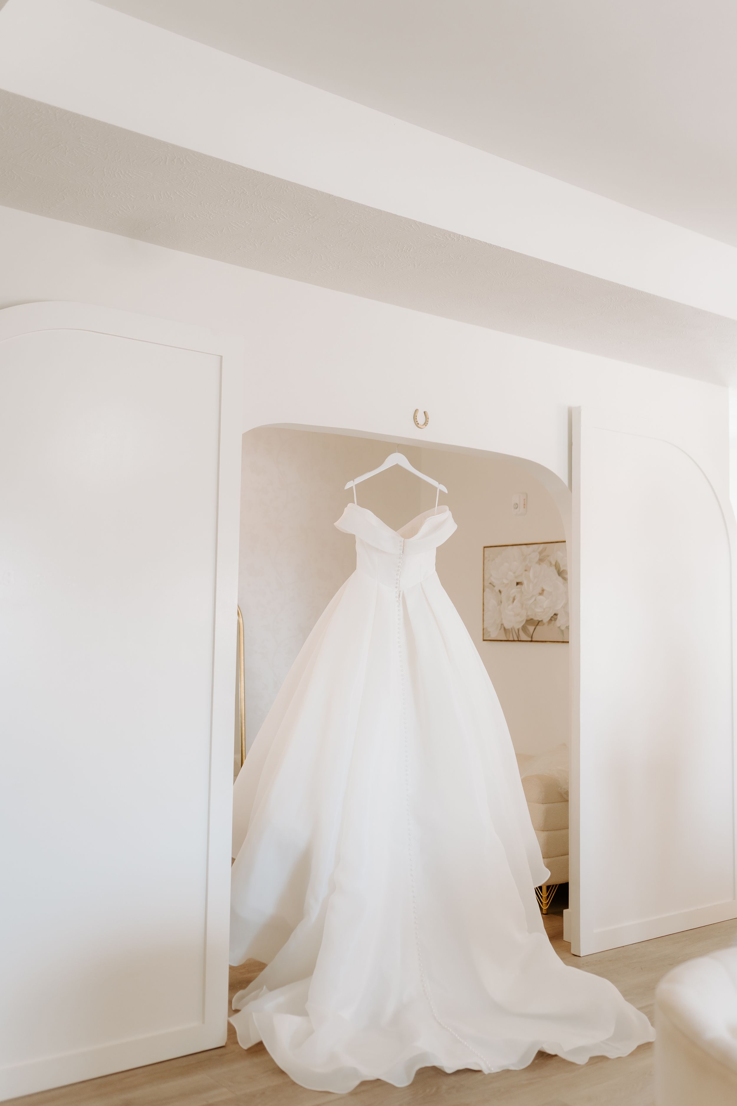 Elegant white off-shoulder ball gown wedding dress hanging on a hanger in a bright, airy bridal suite.