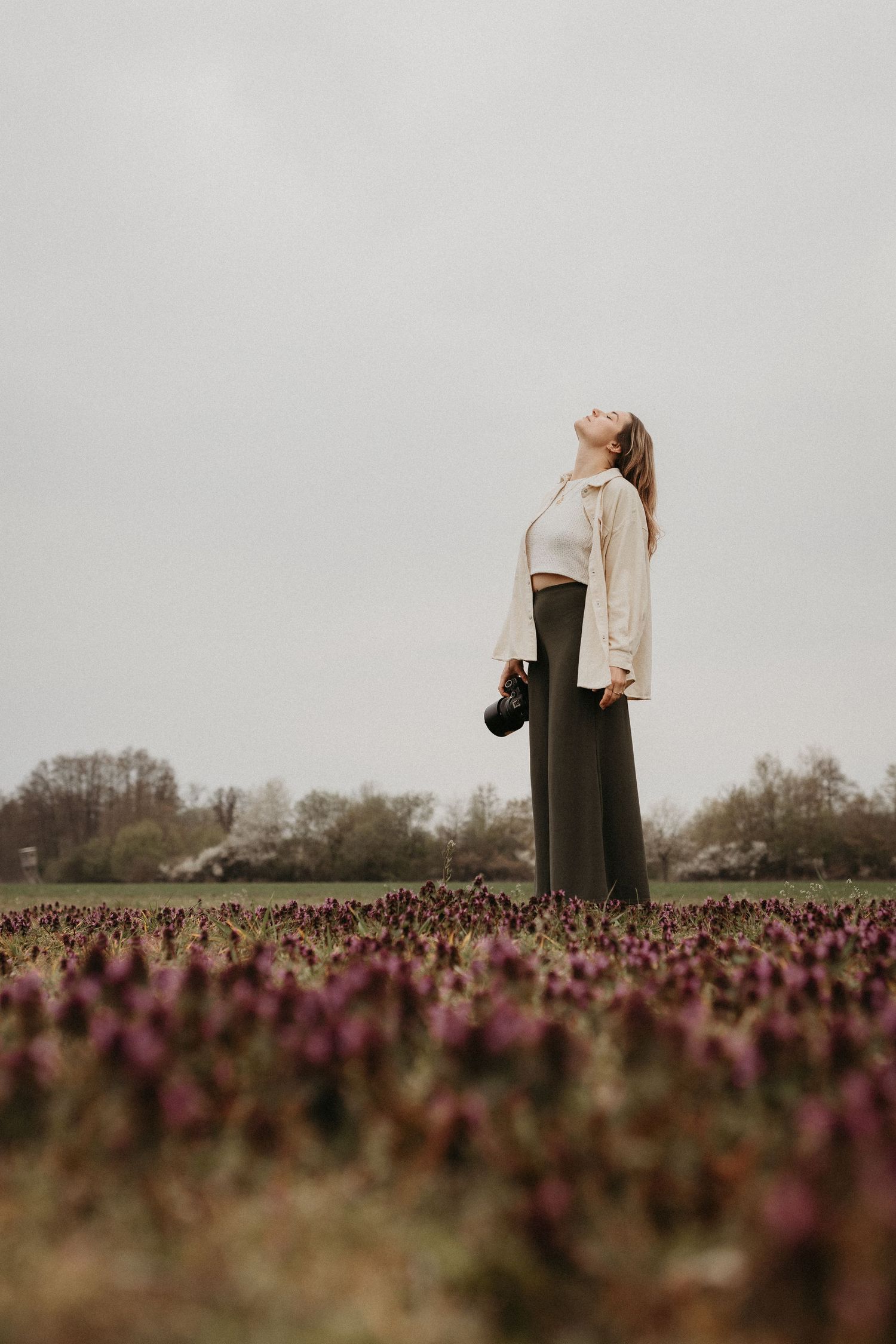 Woman standing in a purple flower field, head tilted back, holding a camera, wearing wide-leg pants and a cream jacket.