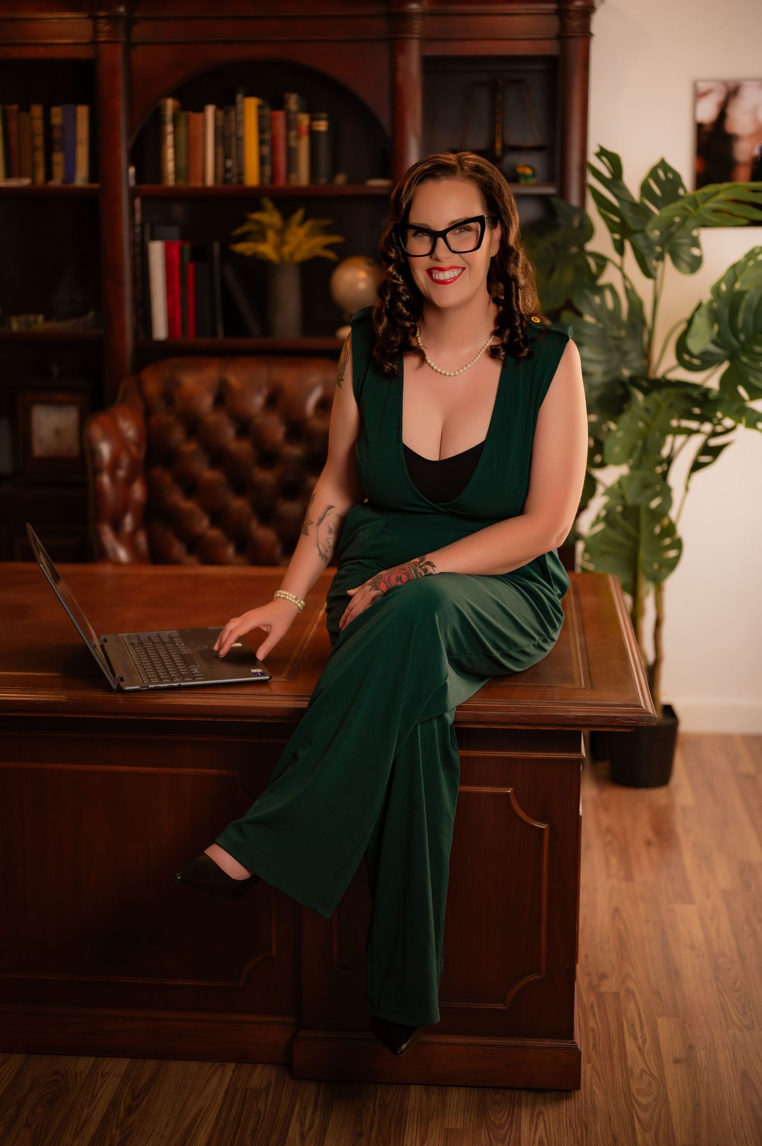 Woman in green jumpsuit sitting on wooden desk with laptop in elegant library office setting.