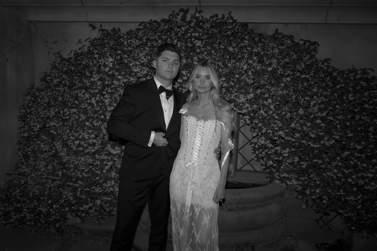 A black and white photo of a couple in formal attire posing in front of a floral wall backdrop at an event.