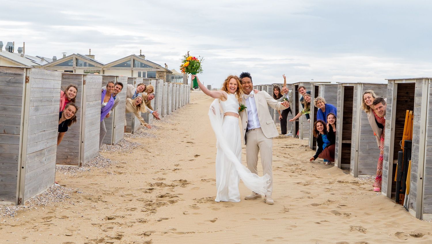 Bruid en bruidegom lopen tussen strandhuisjes door, terwijl lachende bruiloftsgasten door de deuren naar buiten gluren.