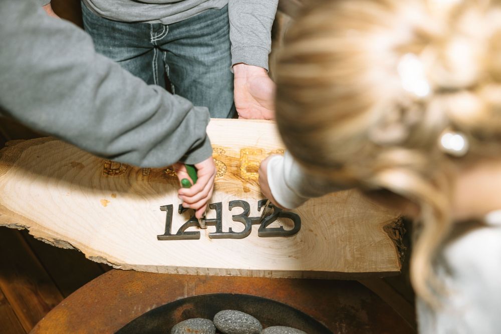 Couple crafting a rustic wooden wedding sign with metal numbers on a live edge wood slab.