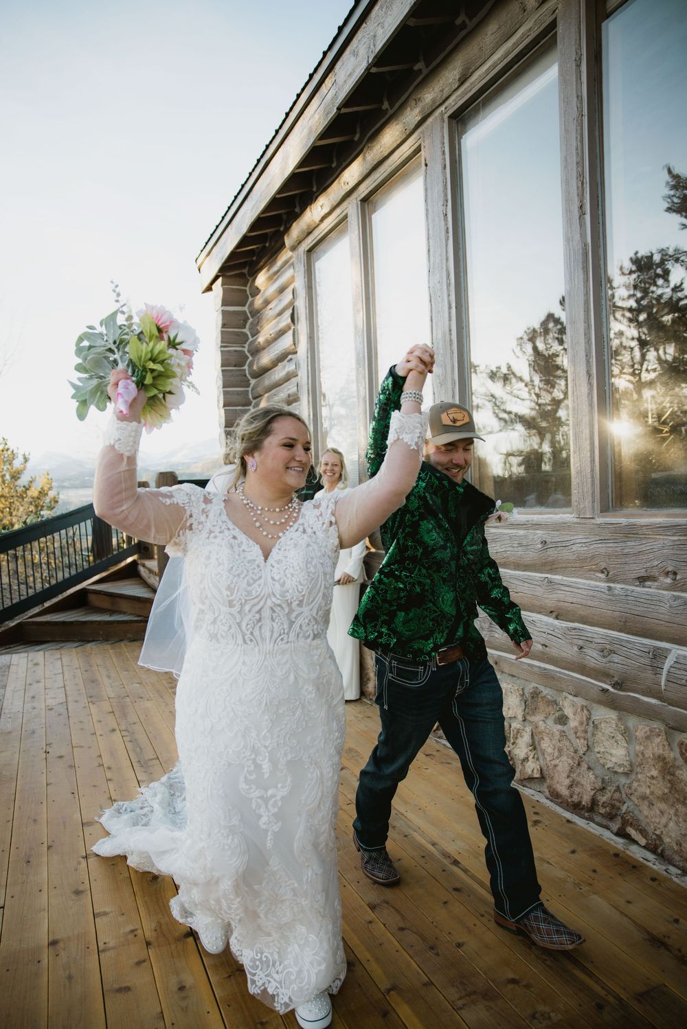 Joyful newlyweds celebrate on wooden deck of rustic venue raising hands with bouquet at sunset.