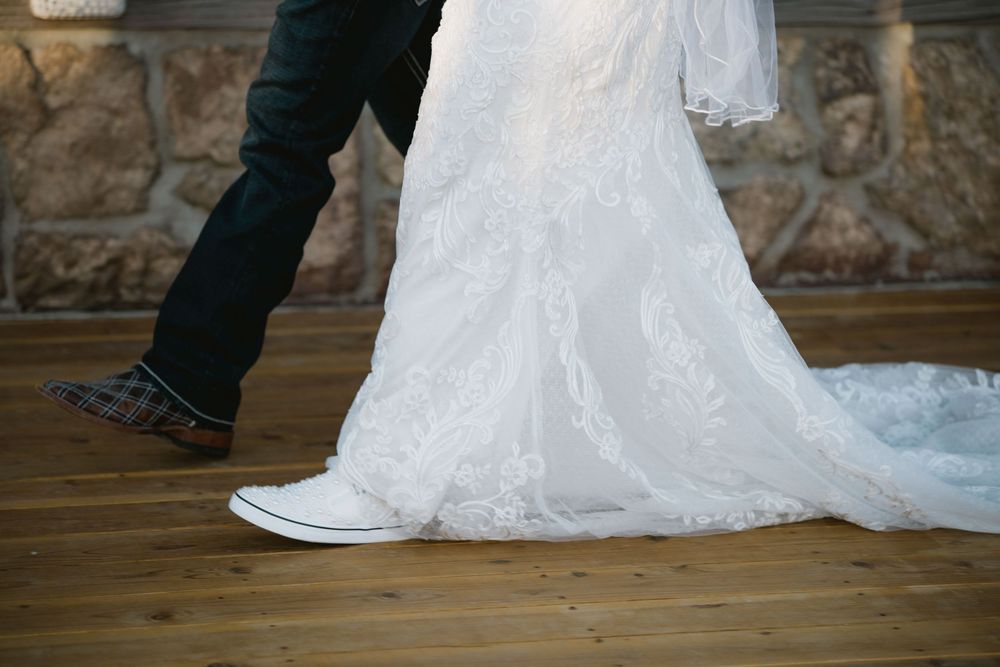 Close-up of bride in white lace wedding gown and groom in dark jeans and Converse sneakers dancing on wooden floor.