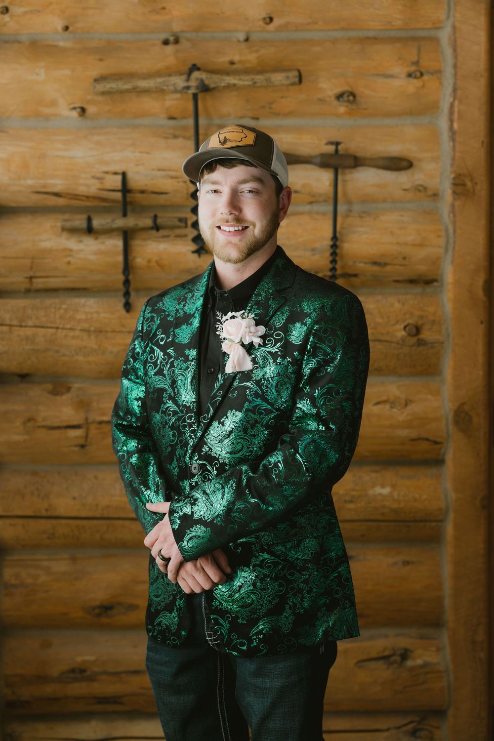 Young man in green floral blazer and trucker hat poses before rustic log wall with boutonniere.