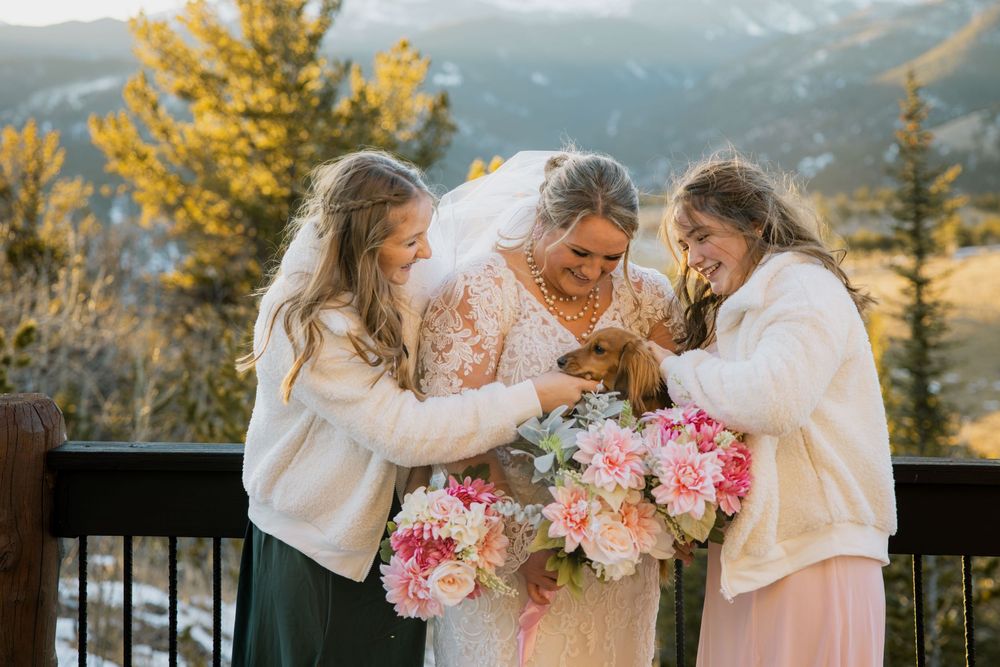 Bride in lace gown with two bridesmaids in white fur jackets holding pink bouquets and a dog on a mountain deck.
