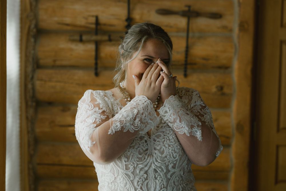 Emotional bride in long-sleeve lace wedding dress covers her face while crying in a rustic wooden cabin.