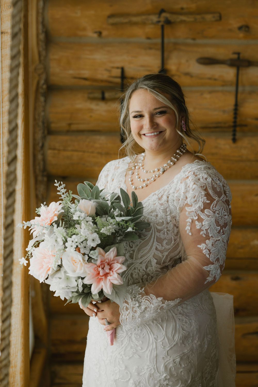 Smiling bride in lace wedding gown holds pink and white bouquet inside rustic log cabin venue.